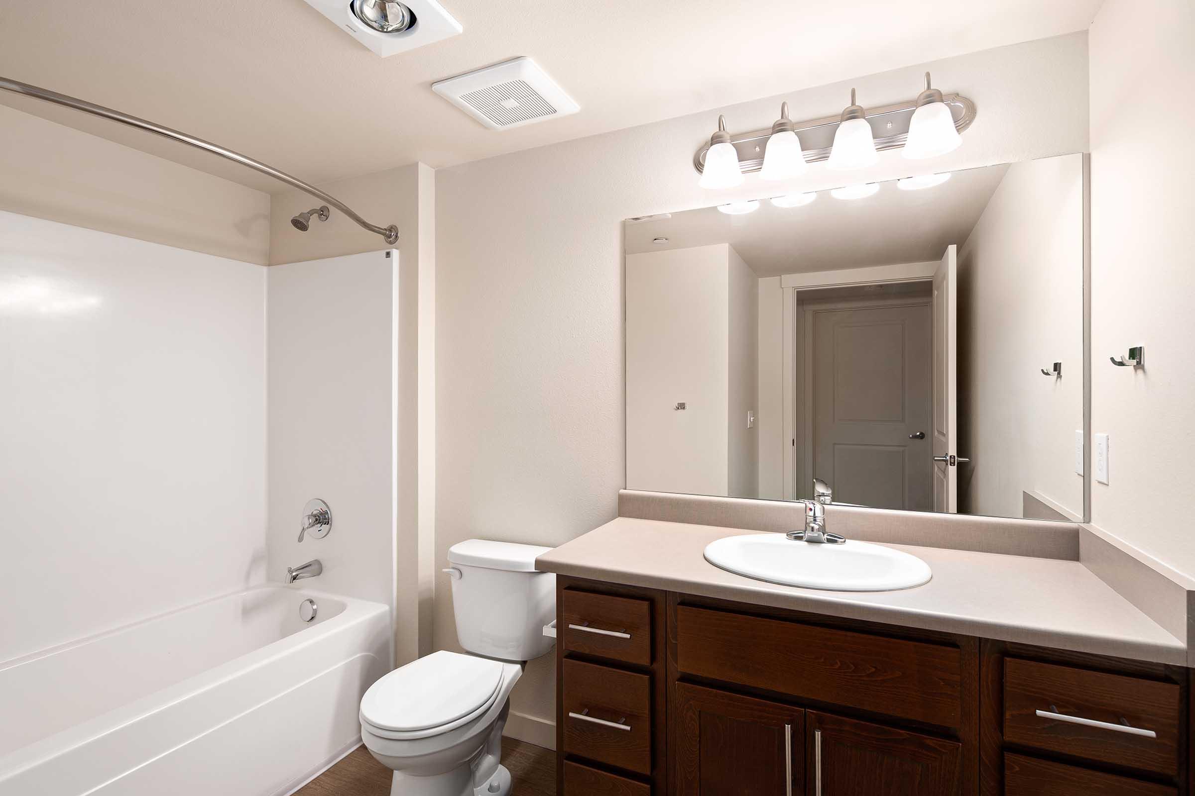 A clean and modern bathroom featuring a bathtub with shower, a toilet, and a single vanity with a sink. The wall above the sink is fitted with lights, and there's a large mirror reflecting the room. The walls are painted in a neutral color, and there's a door leading to another room in the background.