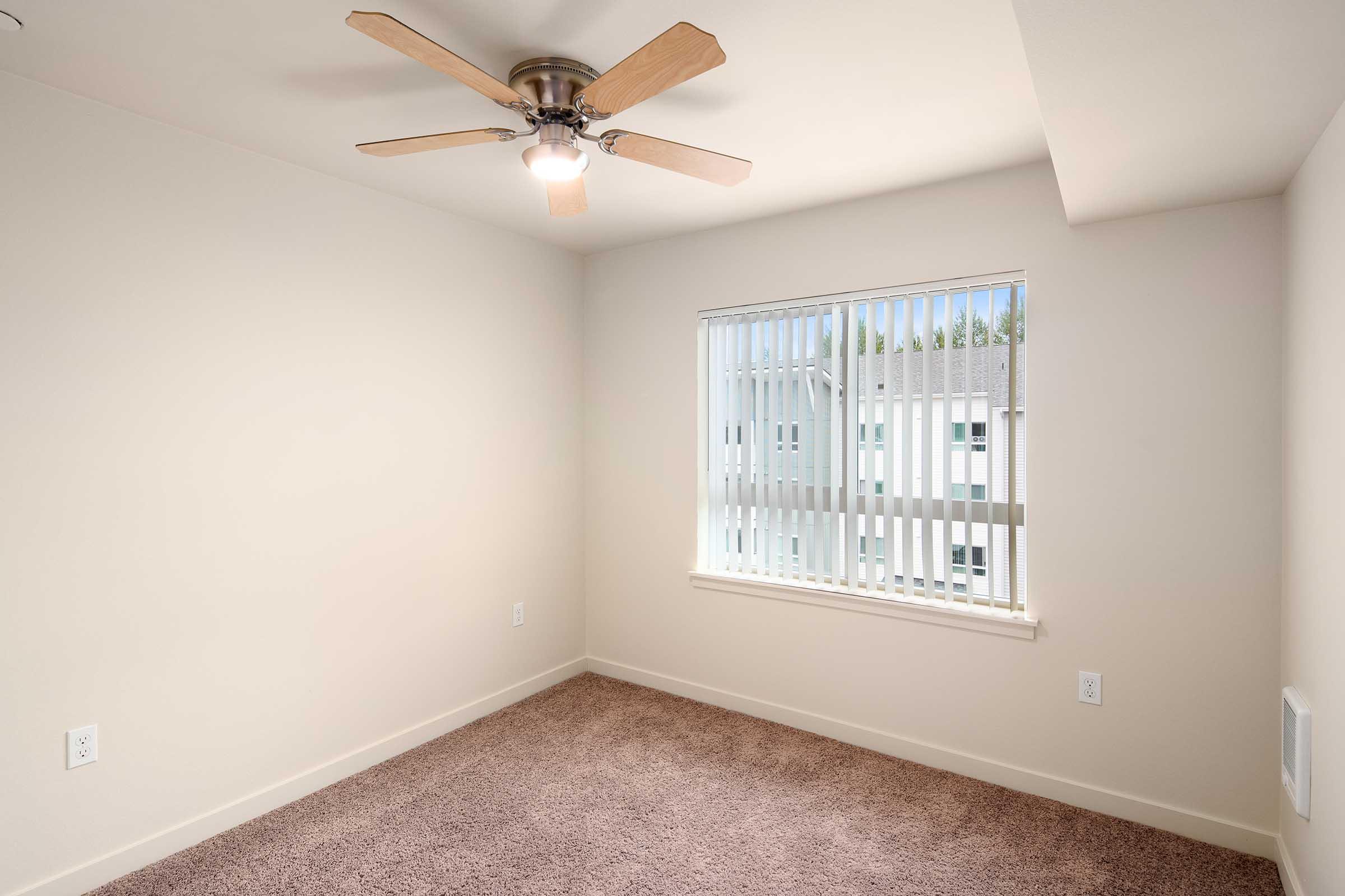 A well-lit room featuring light beige walls, a tan carpeted floor, and a ceiling fan with wooden blades. A window with vertical blinds allows natural light to enter, providing a view of another building outside. The room is empty, creating a spacious and open feel.
