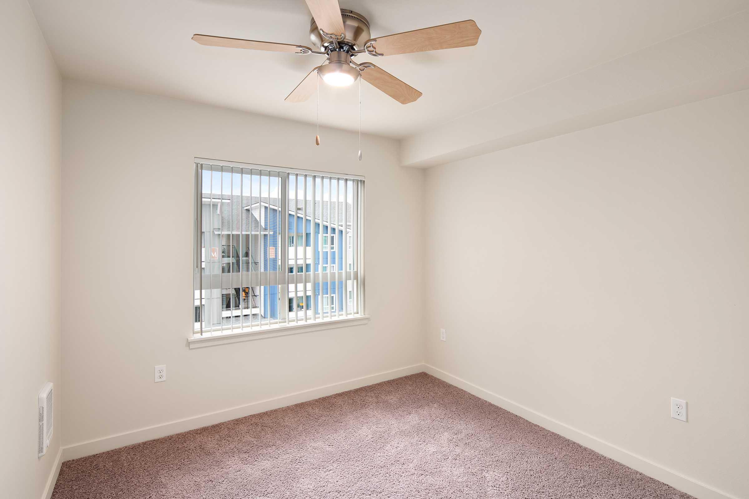 An empty room with light beige walls and a ceiling fan, featuring a window with vertical blinds that lets in natural light. The floor is covered in soft, light brown carpet, and the room has no furniture, creating a clean and minimalistic appearance.