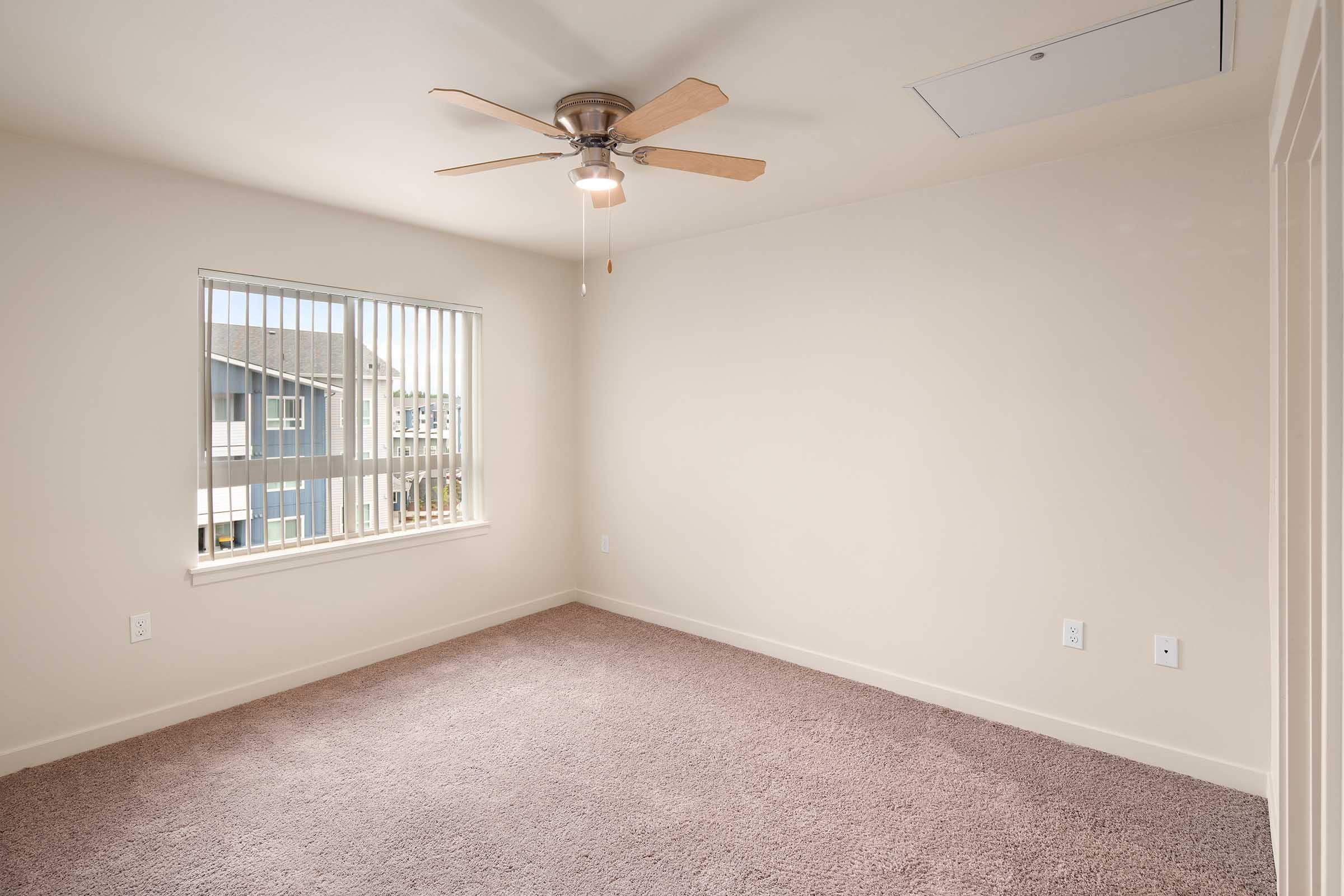 A light, empty room with beige walls and carpeted floor, featuring a ceiling fan with wooden blades. There is a window with vertical blinds allowing natural light, and the room is clean and spacious, suitable for various uses.