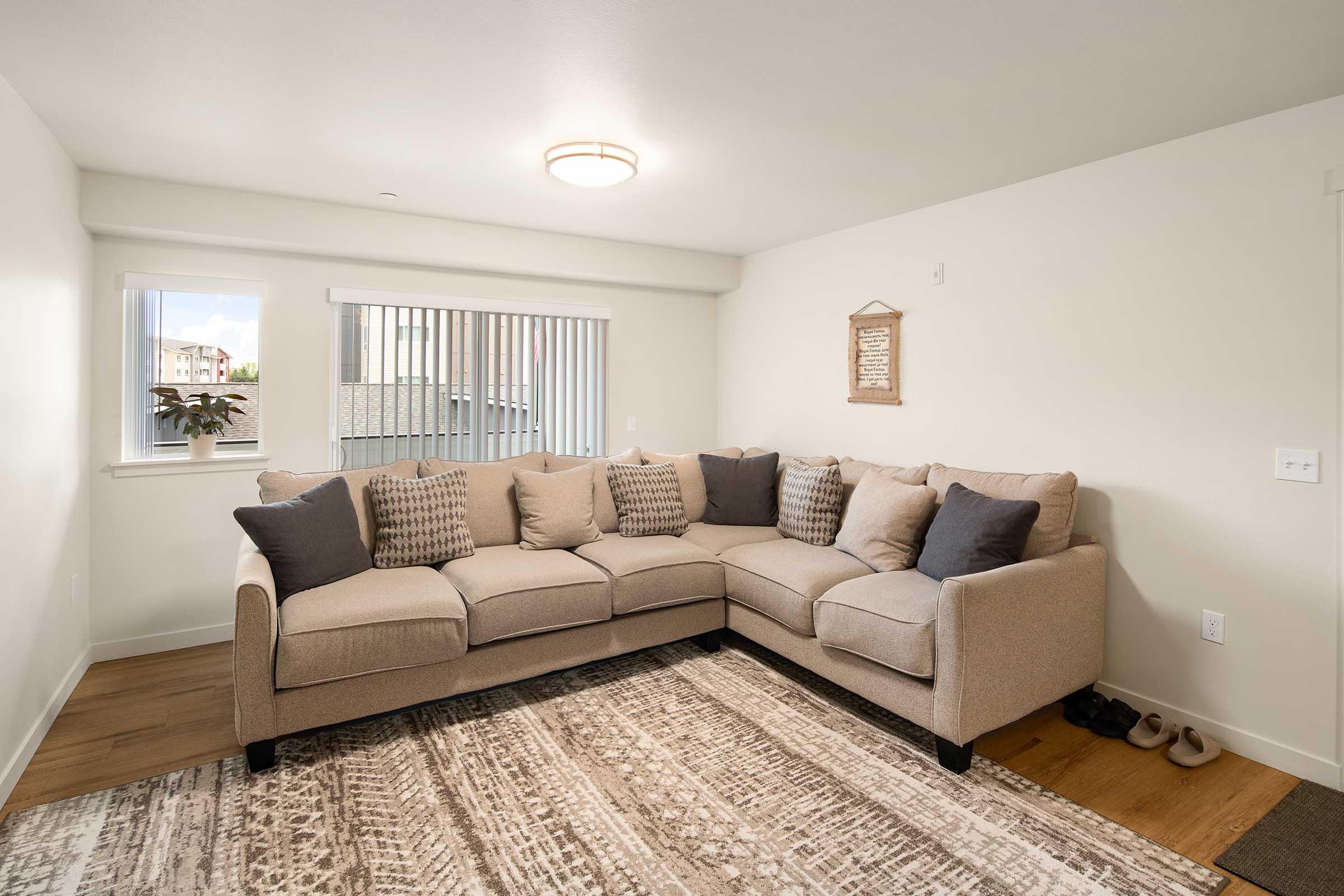 A cozy living room featuring a beige sectional sofa adorned with decorative pillows, positioned on a patterned area rug. Two windows let in natural light, and a small plant is visible. A wall hanging adds a touch of decor, while a pair of slippers can be seen on the floor.