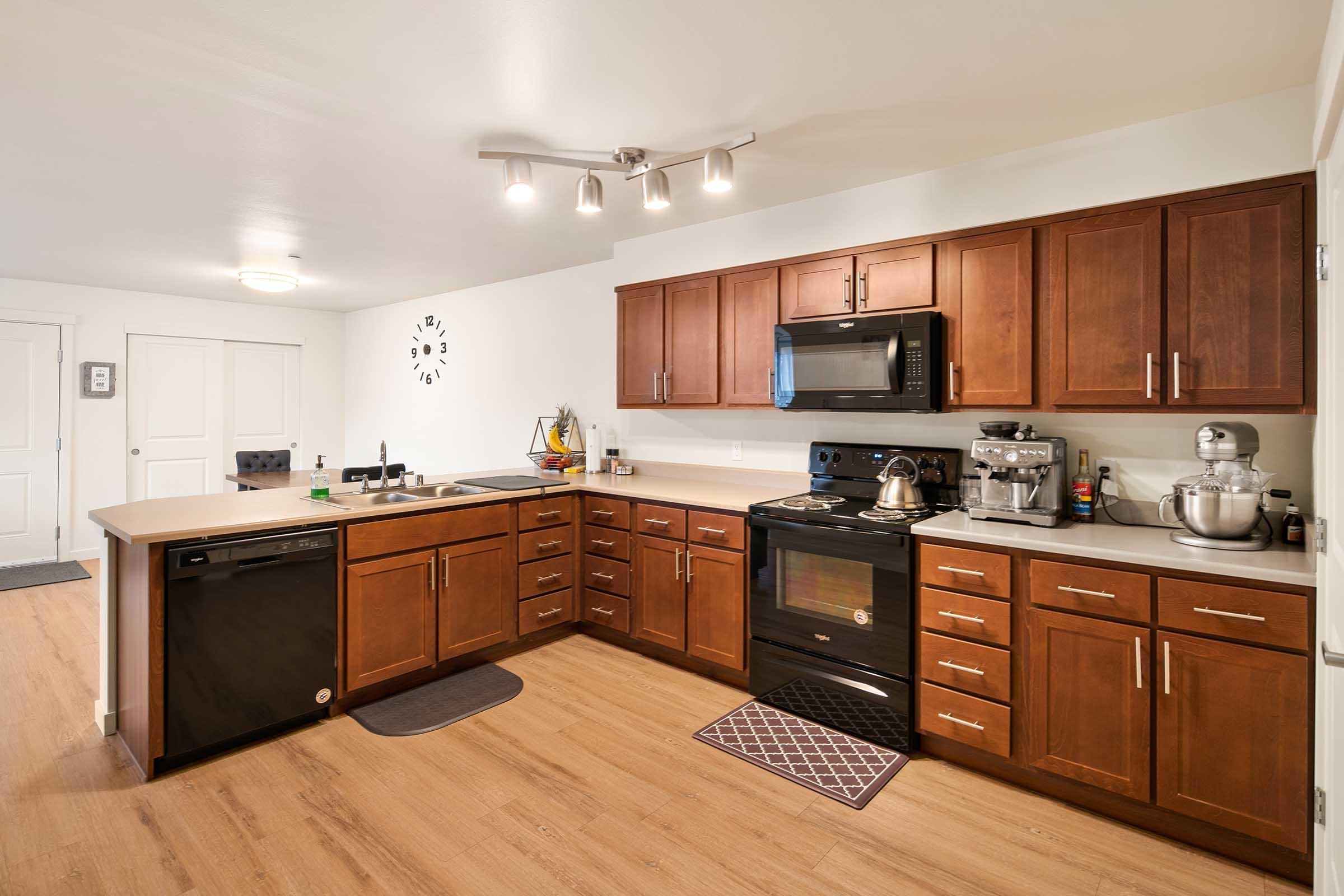 Modern kitchen featuring dark wood cabinets, a countertop with a sink, and stainless steel appliances including a microwave, stove, and dishwasher. The floor is light wood, and there are decorative elements like a clock on the wall. Natural light enhances the warm ambiance.