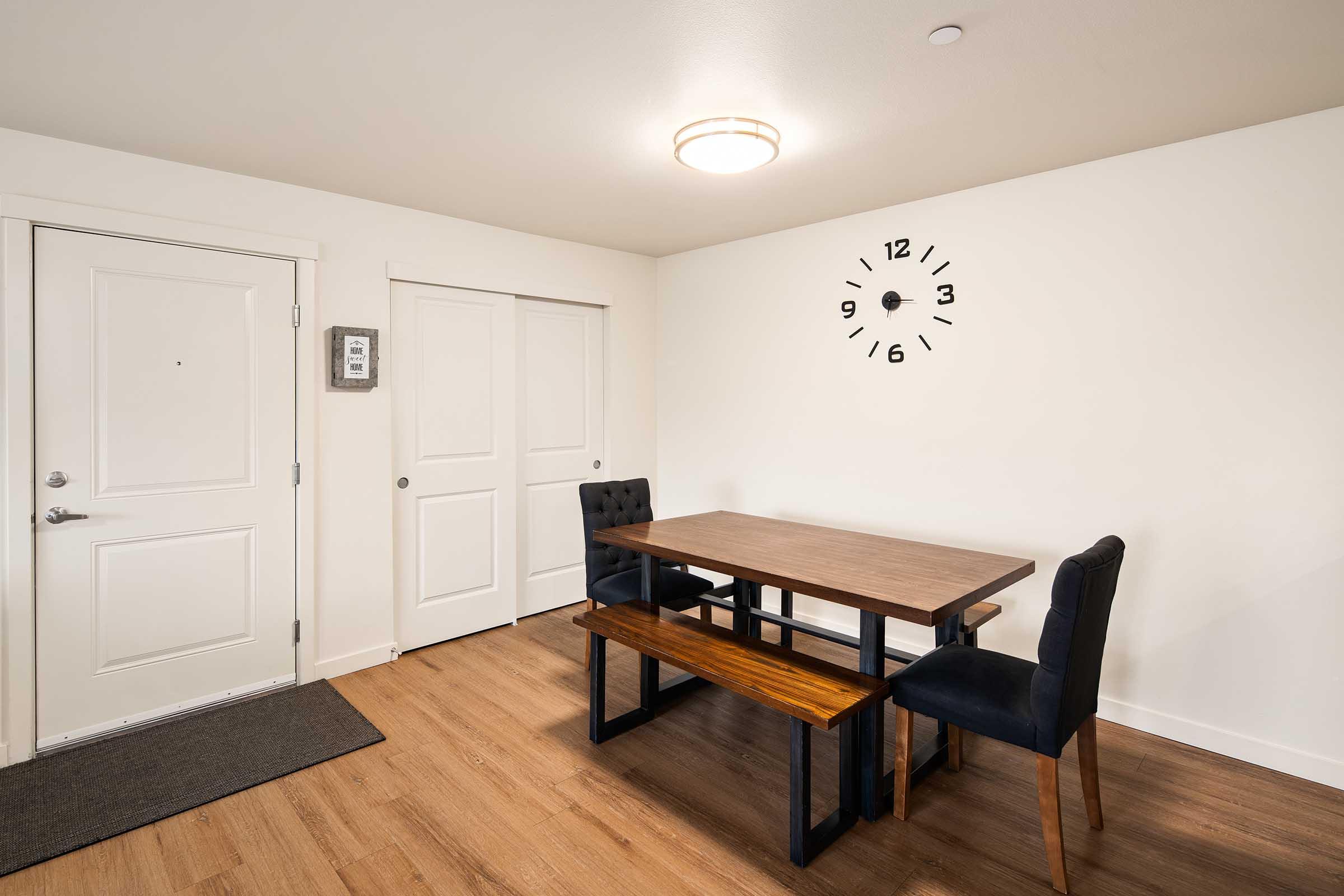 A simple interior space featuring a wooden dining table with a bench and two black chairs. There is a wall clock above the table, showing the time. The entrance door is visible on the left, along with a white closet door. The flooring is wood, and the walls are painted white, creating a modern, minimalist look.