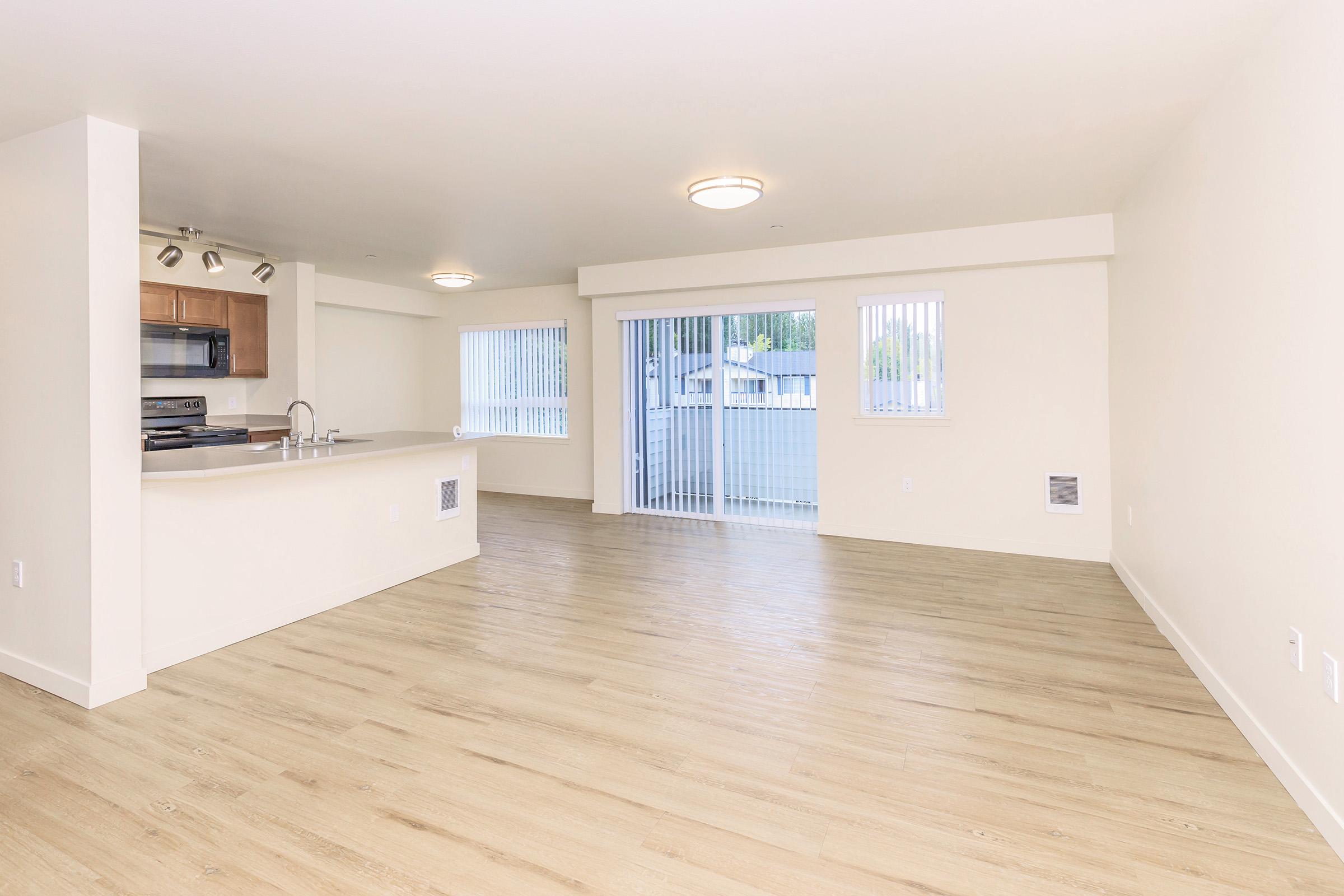 Spacious empty living area with light-colored walls and hardwood flooring. The kitchen features a bar counter and modern appliances. Large sliding glass doors lead to a patio, allowing natural light to enter. Vertical blinds cover the windows, creating a bright and airy atmosphere.