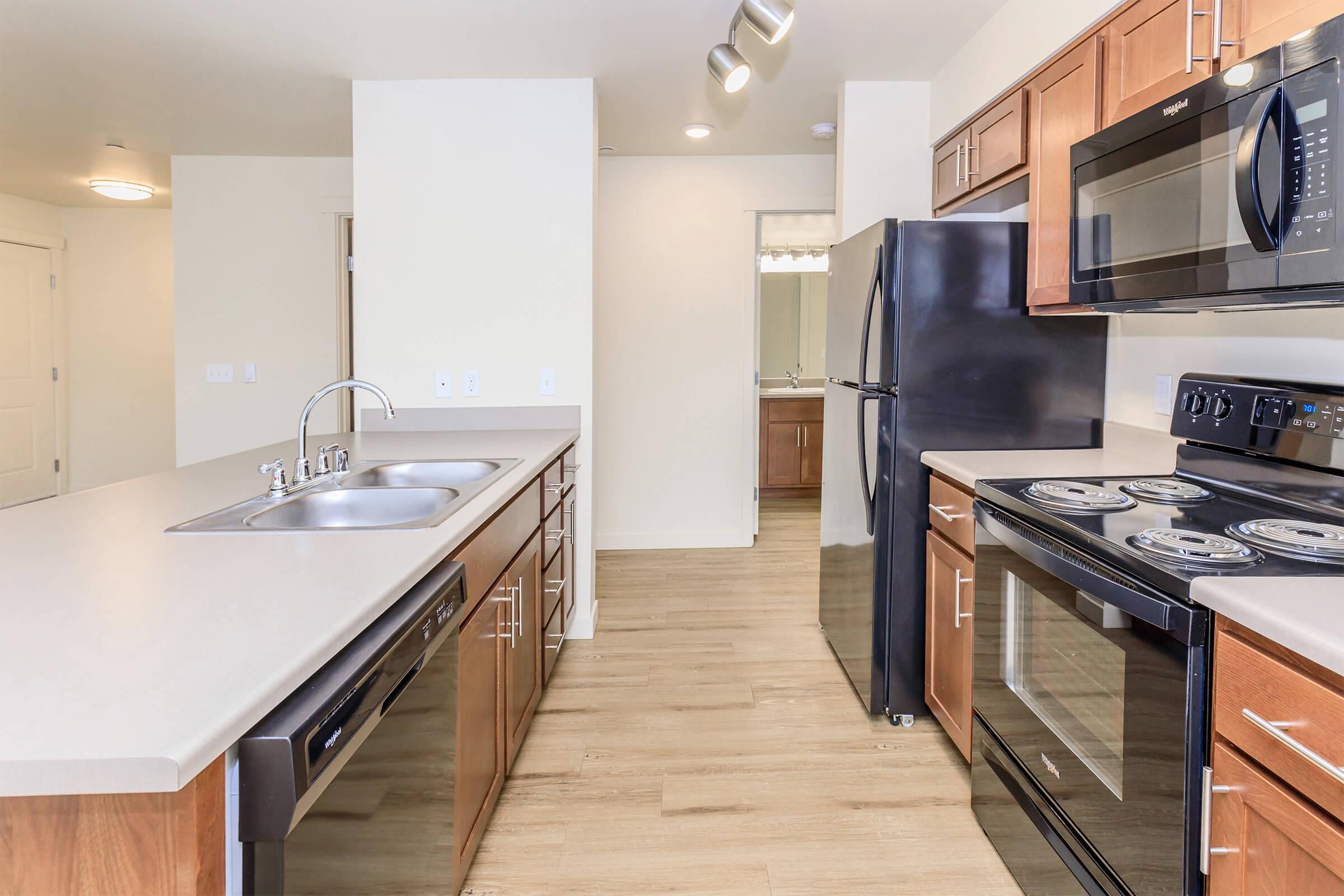 Modern kitchen featuring wood cabinetry, stainless steel appliances including a microwave, oven, and dishwasher. A large island with a double sink and sleek countertops. The open layout connects to adjacent rooms, with a doorway leading to a bathroom in the background.