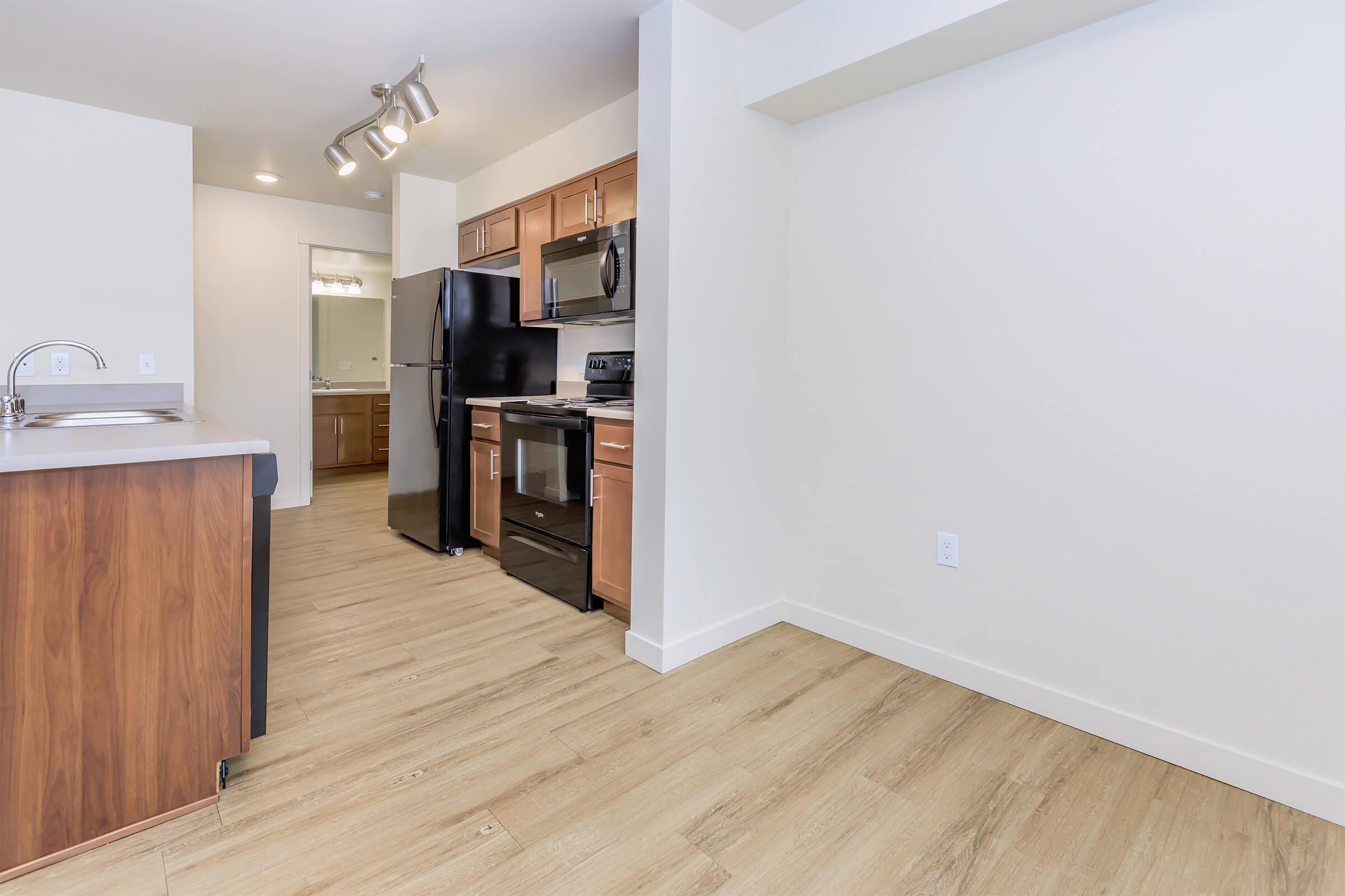 Modern kitchen featuring dark wood cabinetry, stainless steel appliances including a refrigerator and stove, and a breakfast bar. Light-colored walls and flooring create a spacious, inviting atmosphere. A doorway leads to an adjacent area, possibly a bathroom or dining space.