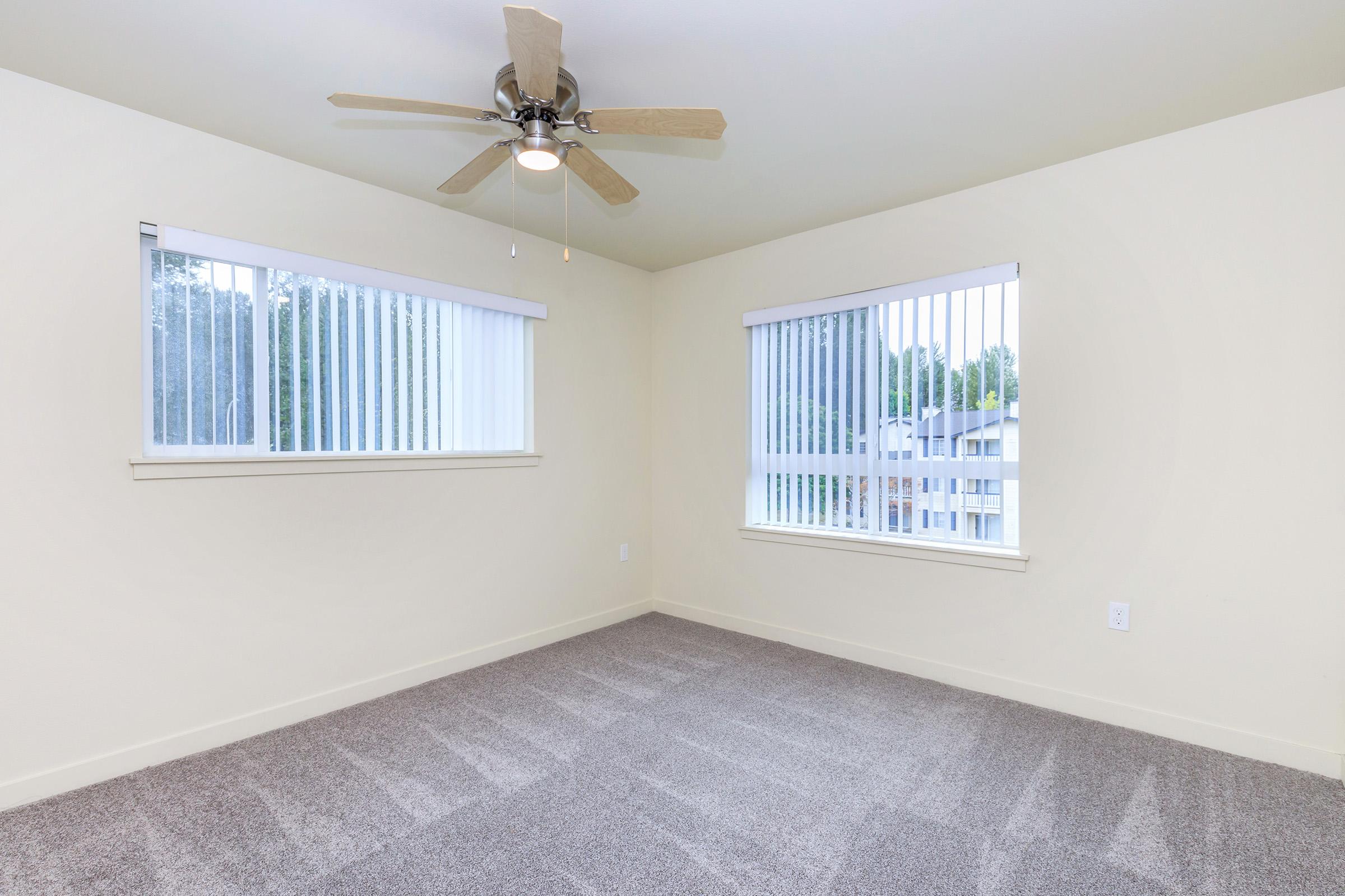 A well-lit, empty room with beige walls and light gray carpeting. It features two windows with vertical blinds, allowing natural light to enter. A ceiling fan is mounted in the center of the room, adding a contemporary touch. The space is clean and inviting, ideal for various uses.