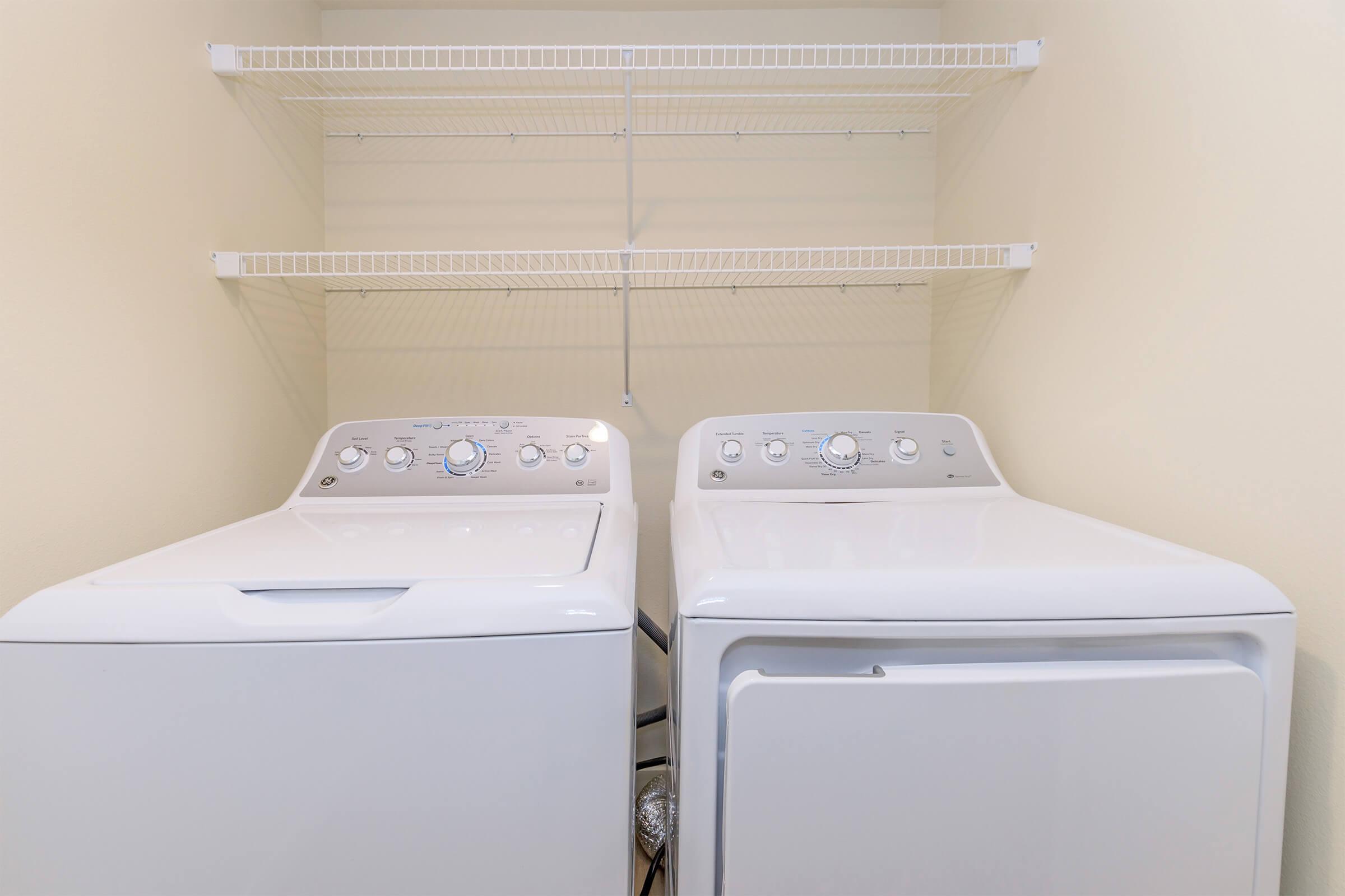 A laundry room featuring a white washing machine and a white dryer side by side, with a beige wall in the background. Above the appliances, there are wire shelves for storage, providing a clean and organized space.