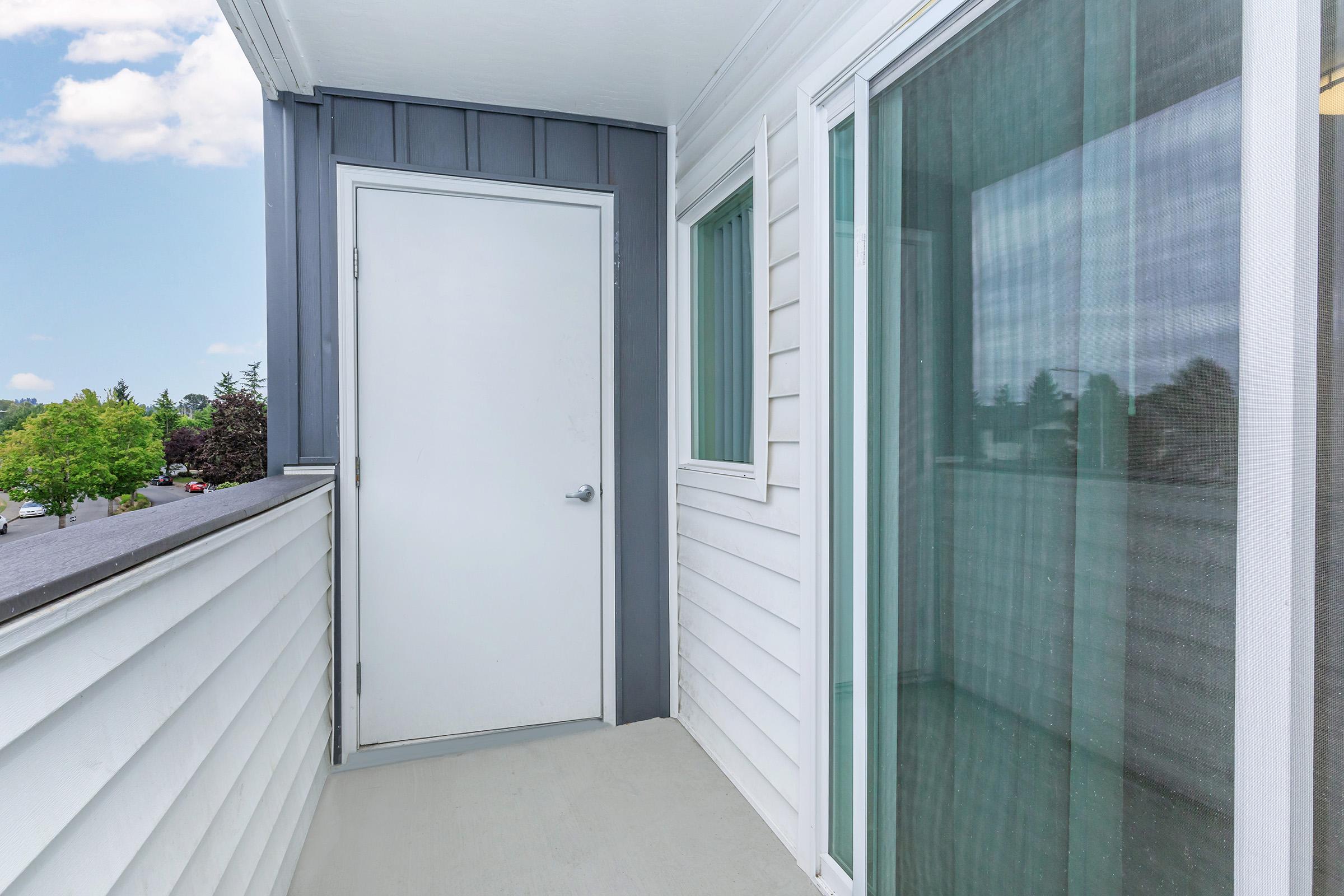 A small balcony featuring a white door on the left, light-colored siding, and a large window with greenish tint on the right. The background includes trees and a blue sky with clouds, creating a serene outdoor atmosphere.