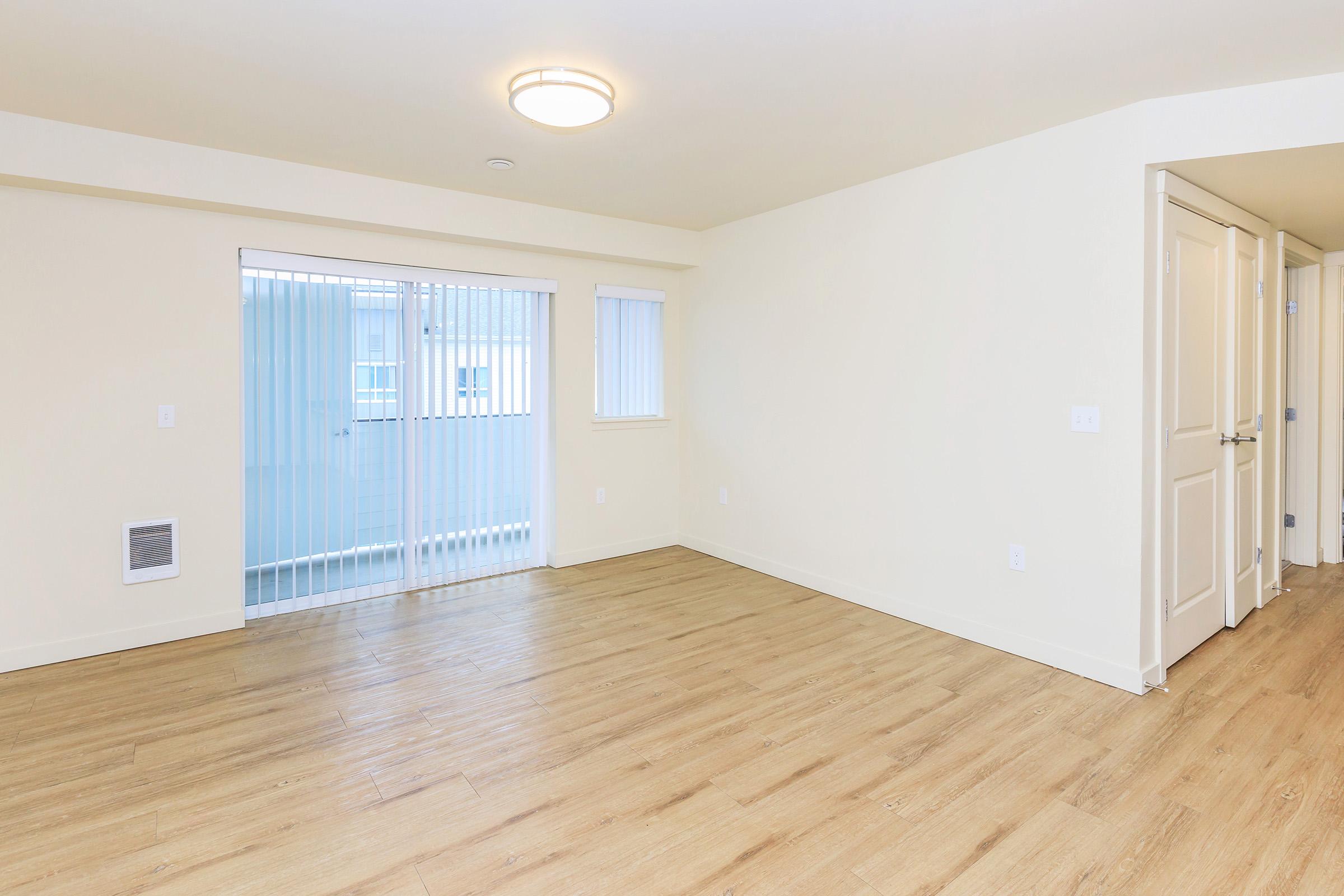 A spacious, empty room with light-colored walls and wood flooring. It features a sliding glass door with vertical blinds that leads to a balcony. The ceiling has a round light fixture, and there are two doorways for entry to other areas. The overall ambiance is bright and airy, suited for furnishing.