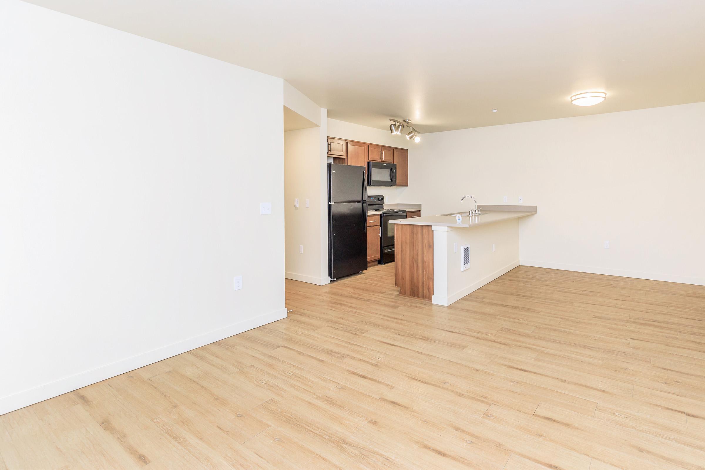 An empty modern living space featuring light-colored walls and light hardwood flooring. The area includes a kitchen with dark cabinets and appliances, a bar counter, and ample open floor space, creating a bright and spacious atmosphere.