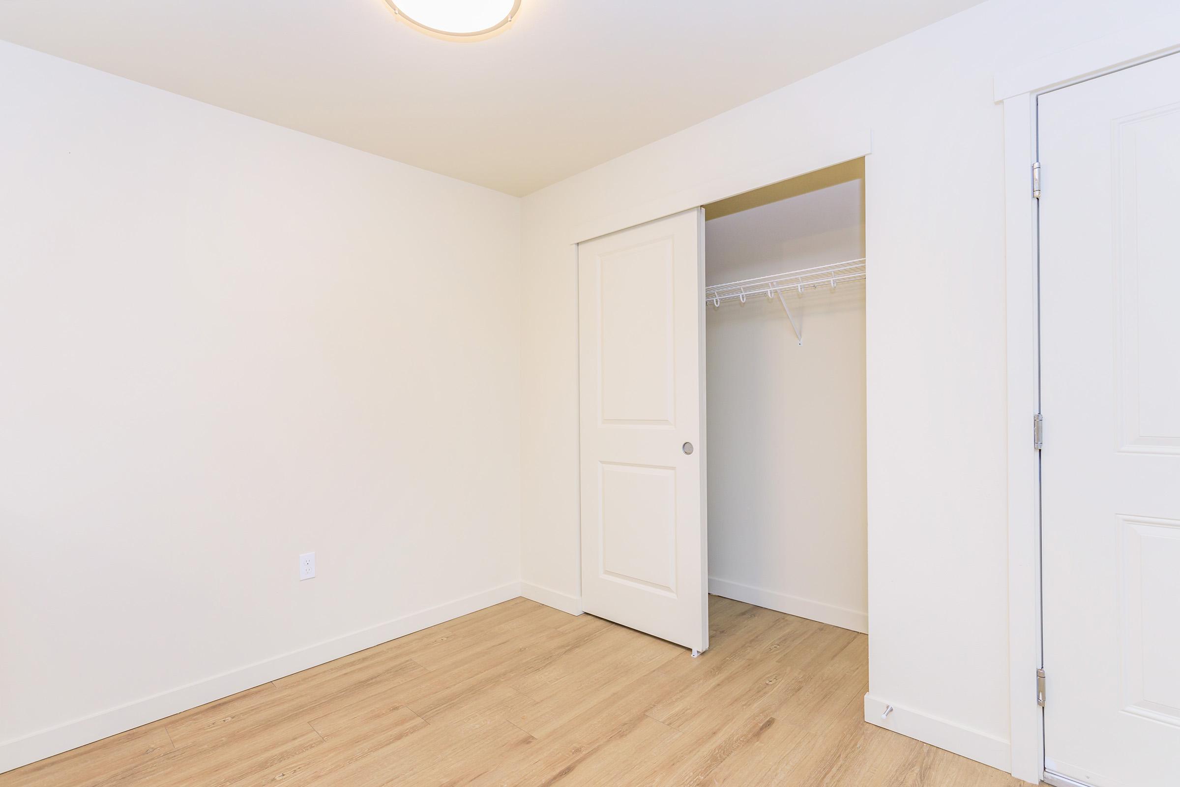 A well-lit, empty room with white walls and a light wood floor. There is a closed door leading to a closet on the left, which has a shelf and hanging space. The ceiling features a simple light fixture, and the overall space appears minimalistic and ready for furnishing.