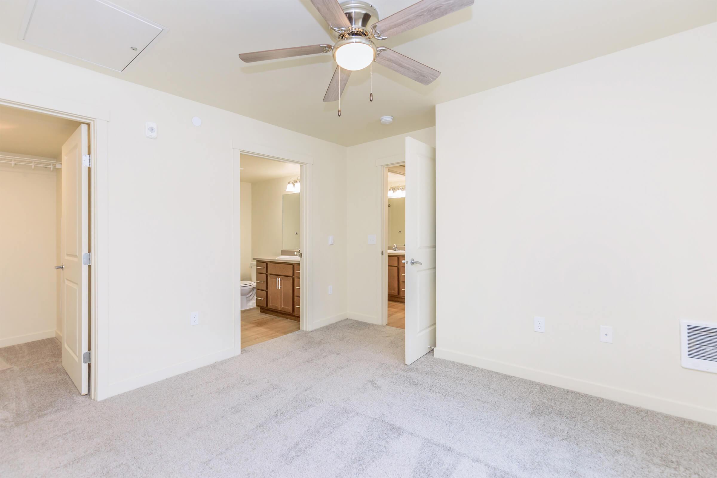 A light-colored room with soft beige walls and gray carpeting. There are two open doorways leading to bathrooms with sinks and mirrors. A ceiling fan is visible above, and the space is well-lit, creating a modern and inviting atmosphere.