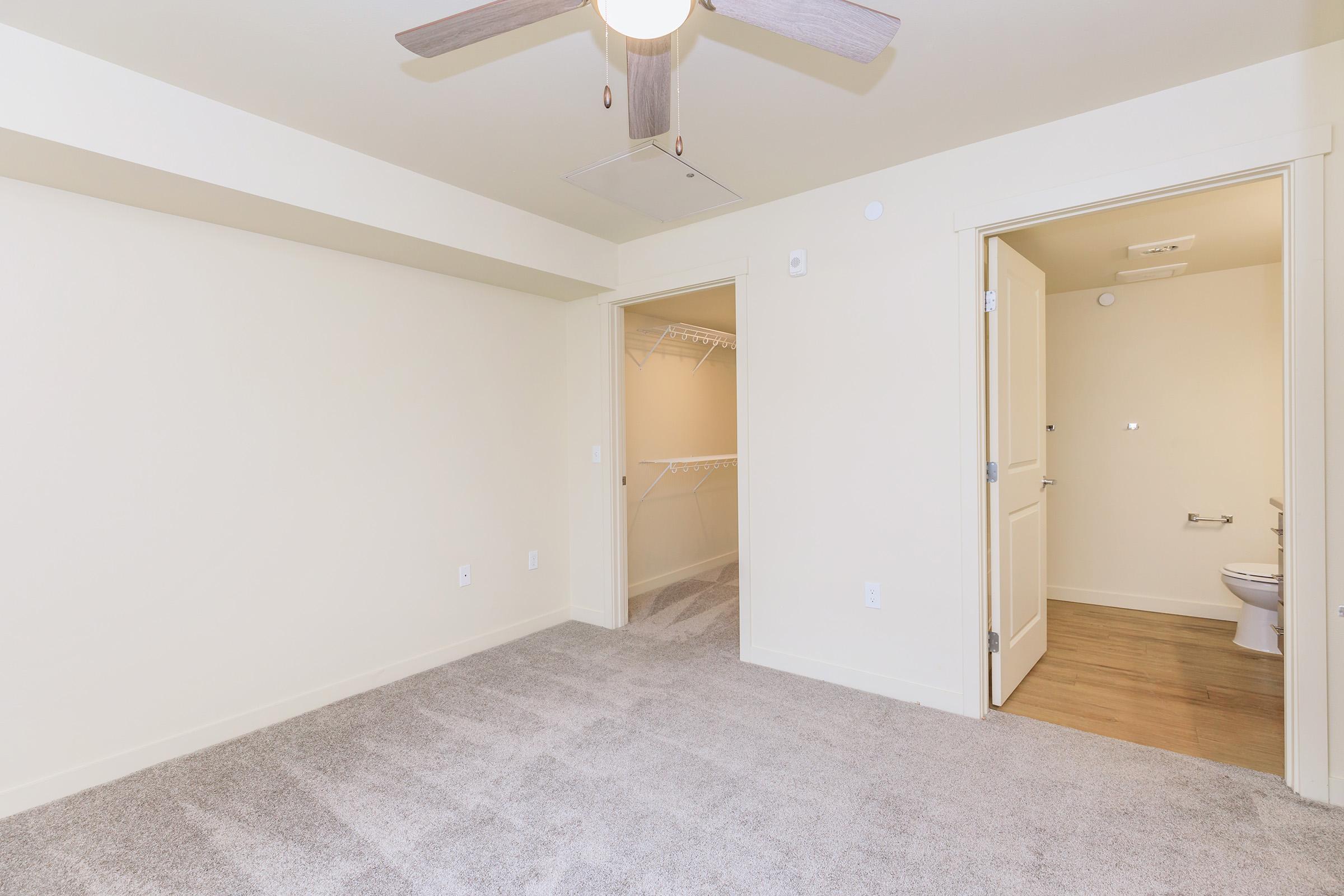 A light-colored room with beige walls and carpet flooring. It features a ceiling fan and an open doorway leading to a bathroom. A second door opens to a spacious closet with a wire shelf. The room is bright and empty, creating a clean and airy feel.
