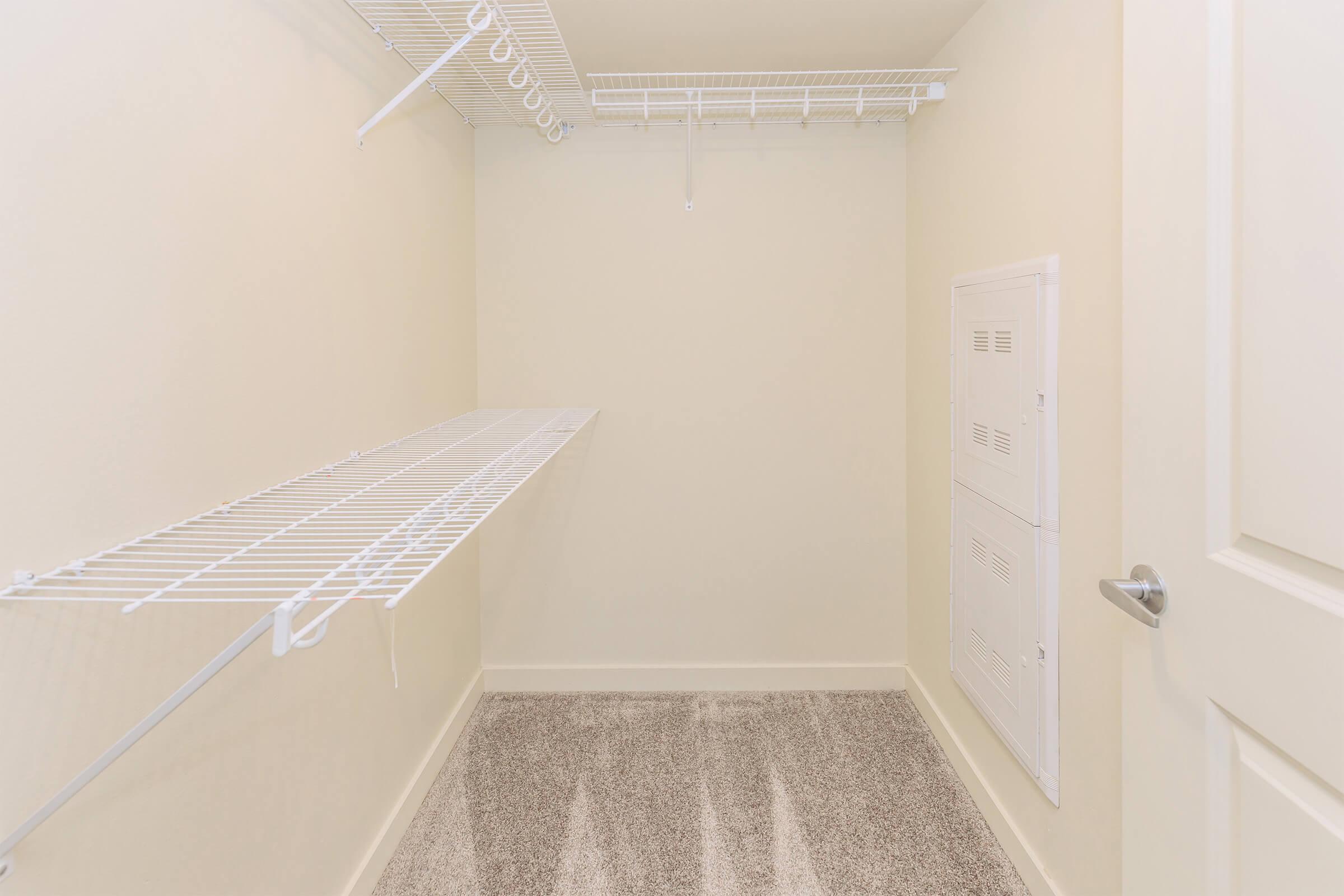 A spacious, empty closet featuring white wire shelving and a beige wall. The room has carpeted flooring and a small utility panel on the side. The shelving has a long ledge for storage, creating a clean and organized look.