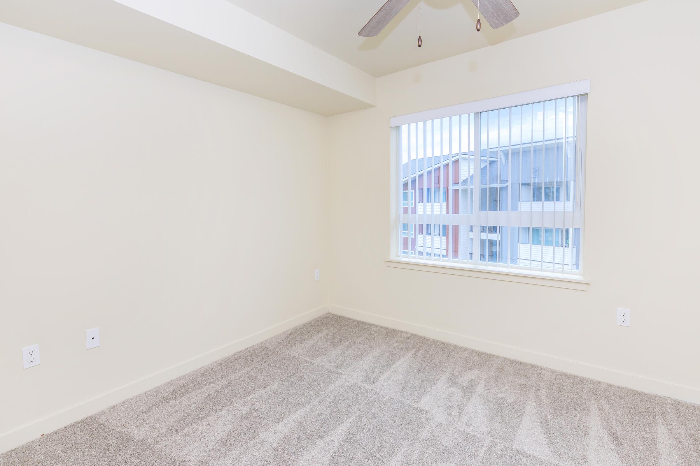 Bright and empty room with beige walls and light gray carpeting. A window with vertical blinds allows natural light to enter, revealing a view of buildings outside. The ceiling features a ceiling fan, adding to the airy feel of the space.