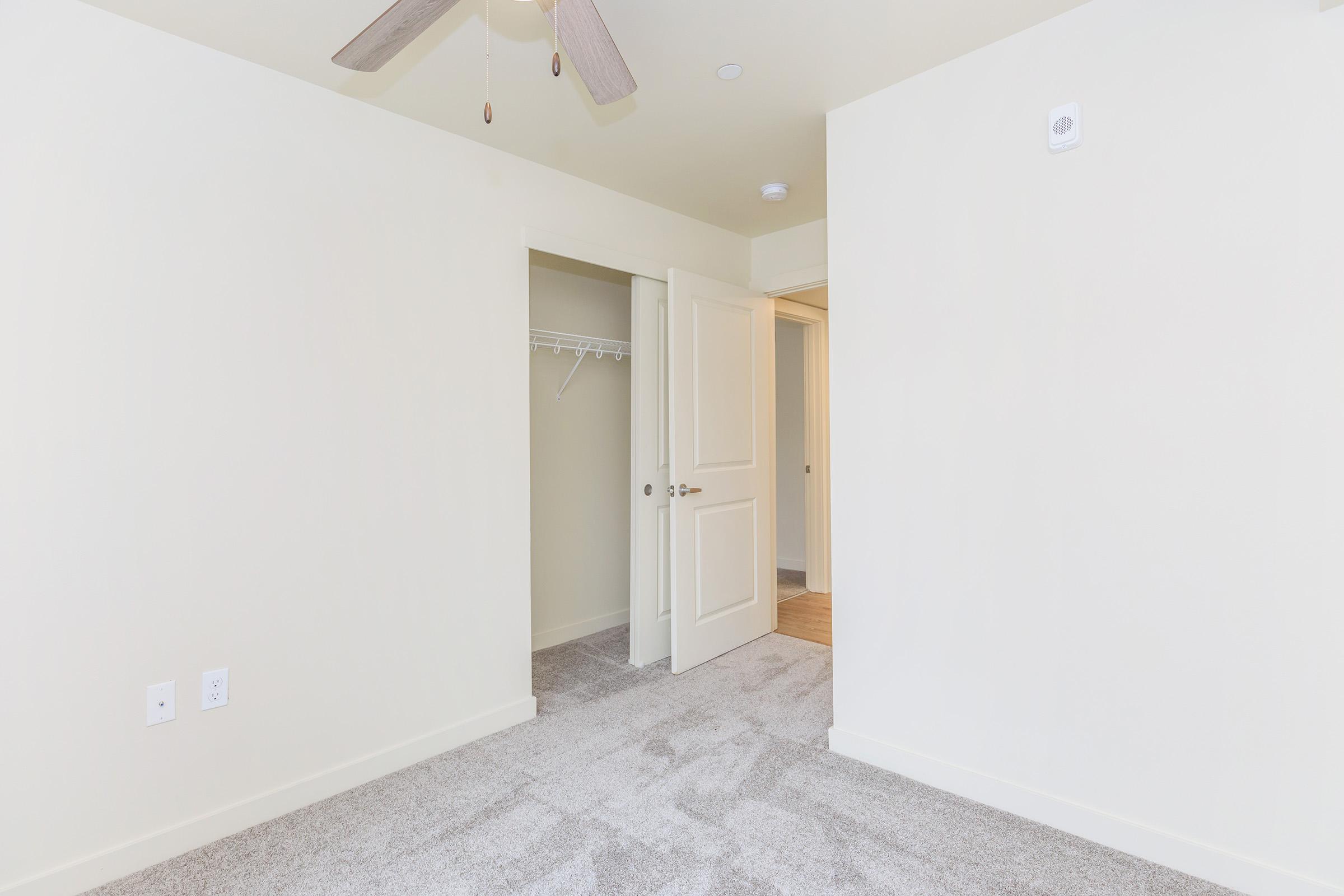A vacant room featuring light-colored walls and a ceiling fan. There's a closet with a door on the left, and another door leading to a hallway at the end of the room. The carpet is plush and beige, creating a neutral and inviting space.