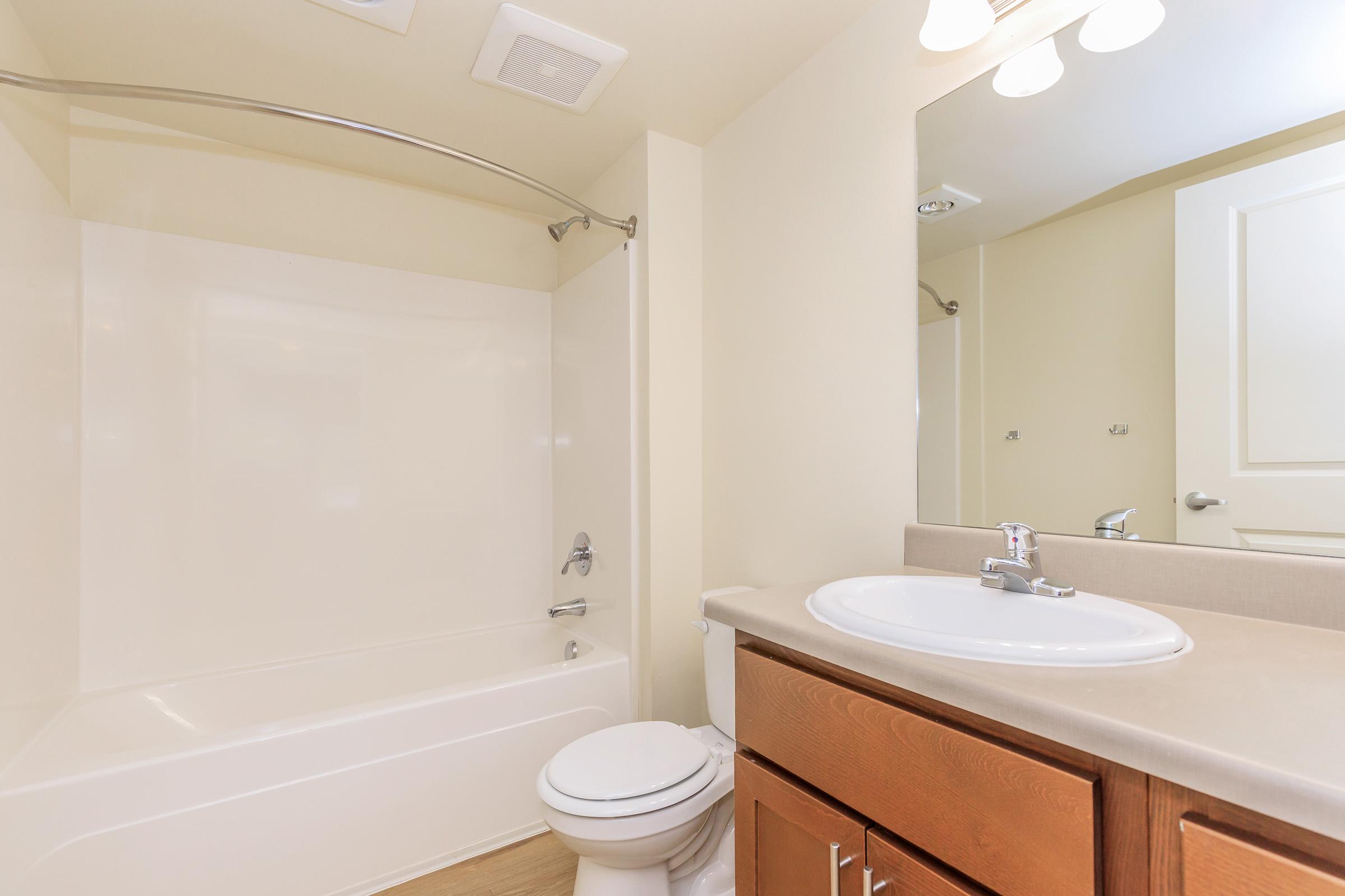 A clean and modern bathroom featuring a bathtub with a curved shower rod, a toilet, and a sink with a wooden vanity. The walls are painted a light color, and there is a mirror above the sink. The lighting is bright, creating a spacious and inviting atmosphere.