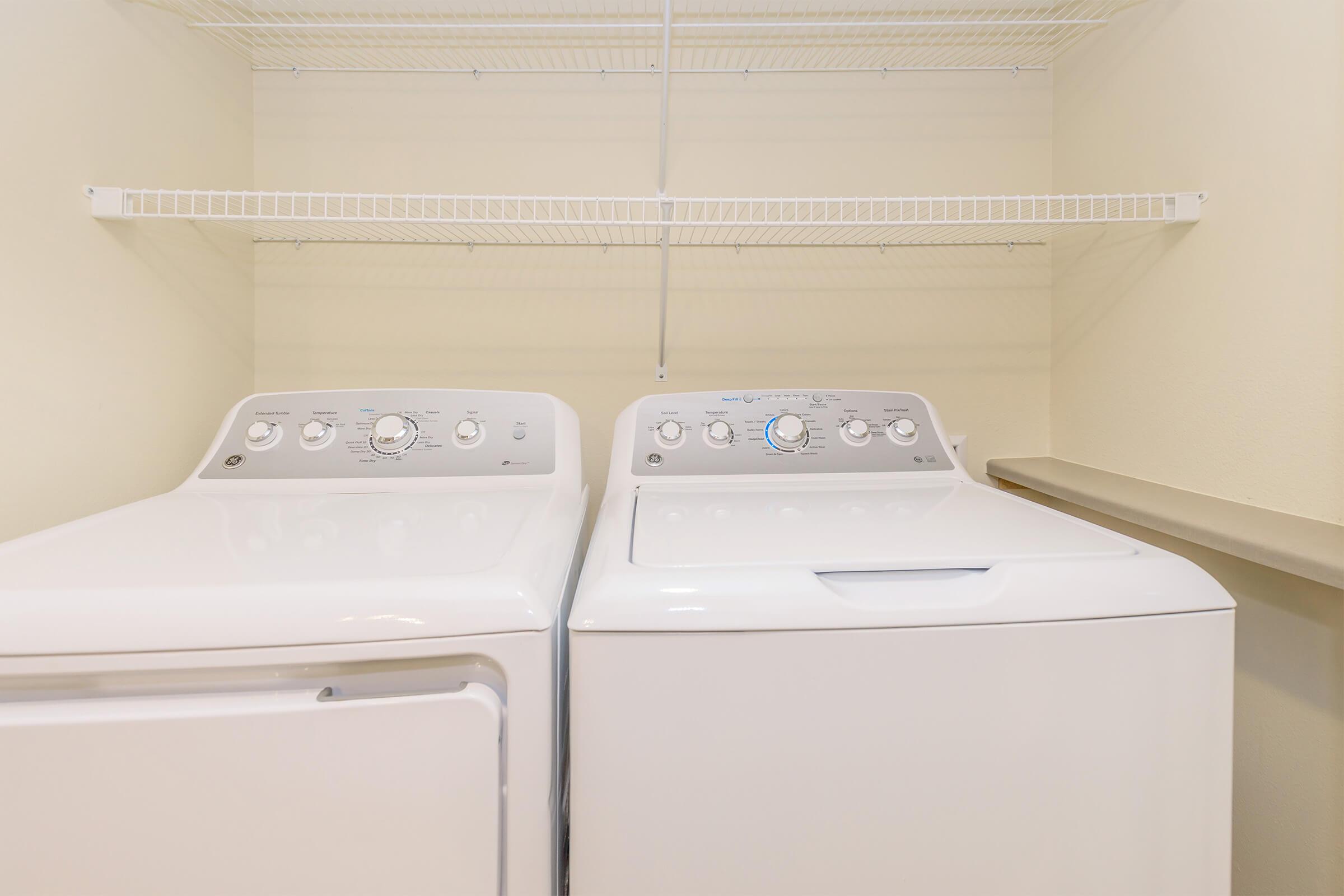 A clean laundry room features a white washer and dryer side by side on a beige floor. Above them, there is a wire shelf for storage. The walls are painted light yellow, creating a bright, airy atmosphere.