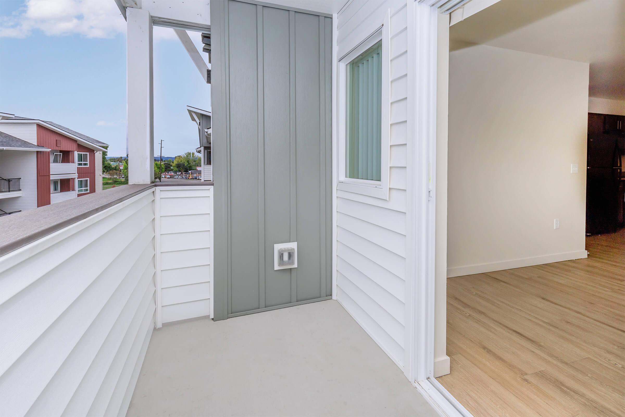 A modern balcony view from an apartment, featuring light-colored walls, a small window, and a door leading inside. The exterior displays a mix of siding textures, with a clear view of surrounding buildings and greenery in the background. The flooring is light wood, enhancing the spacious feel.