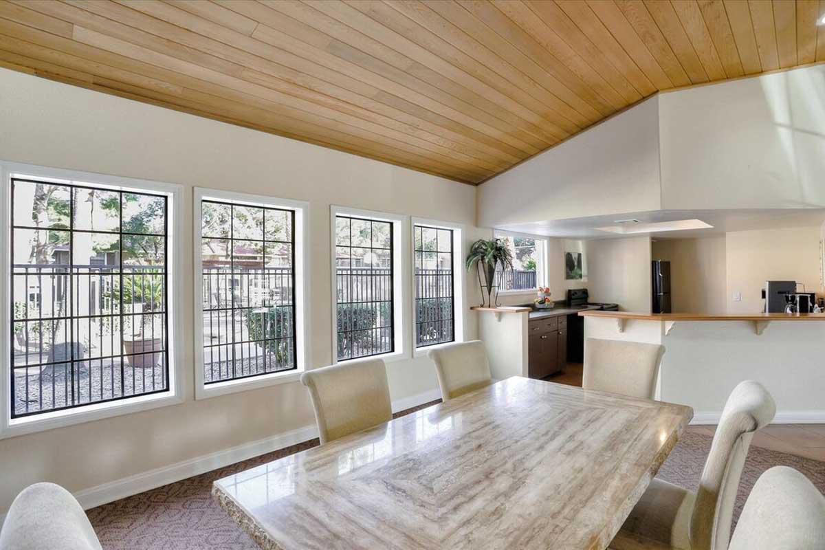 Bright and spacious dining area featuring a large marble table surrounded by beige chairs. Large windows allow natural light and offer views of an outdoor patio and greenery. The room has a warm wooden ceiling and leads into a modern kitchen area.