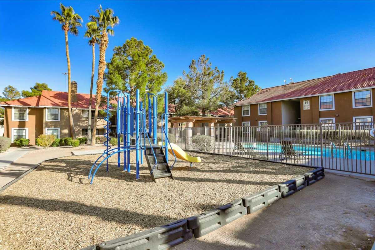 Playground equipment featuring slides and climbing structures in a residential complex, surrounded by palm trees. A swimming pool is visible in the background, with two-story buildings adjacent to the area. The scene is sunny and well-maintained, suggesting a recreational space for families.