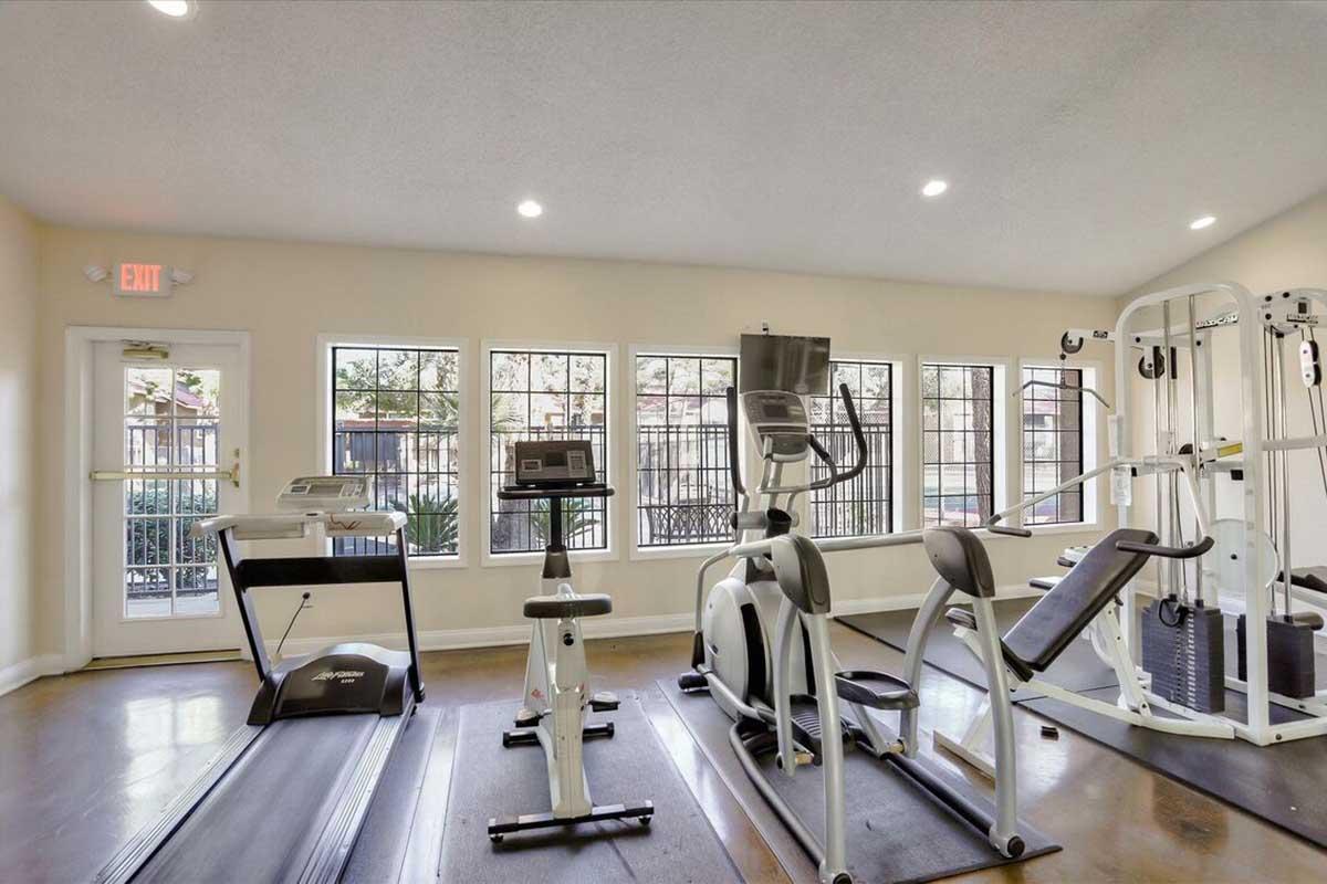 A bright and spacious gym area featuring fitness equipment such as treadmills, elliptical machines, and weight training apparatus. Large windows allow natural light to fill the room, and the space is well-maintained with a clean, modern aesthetic.