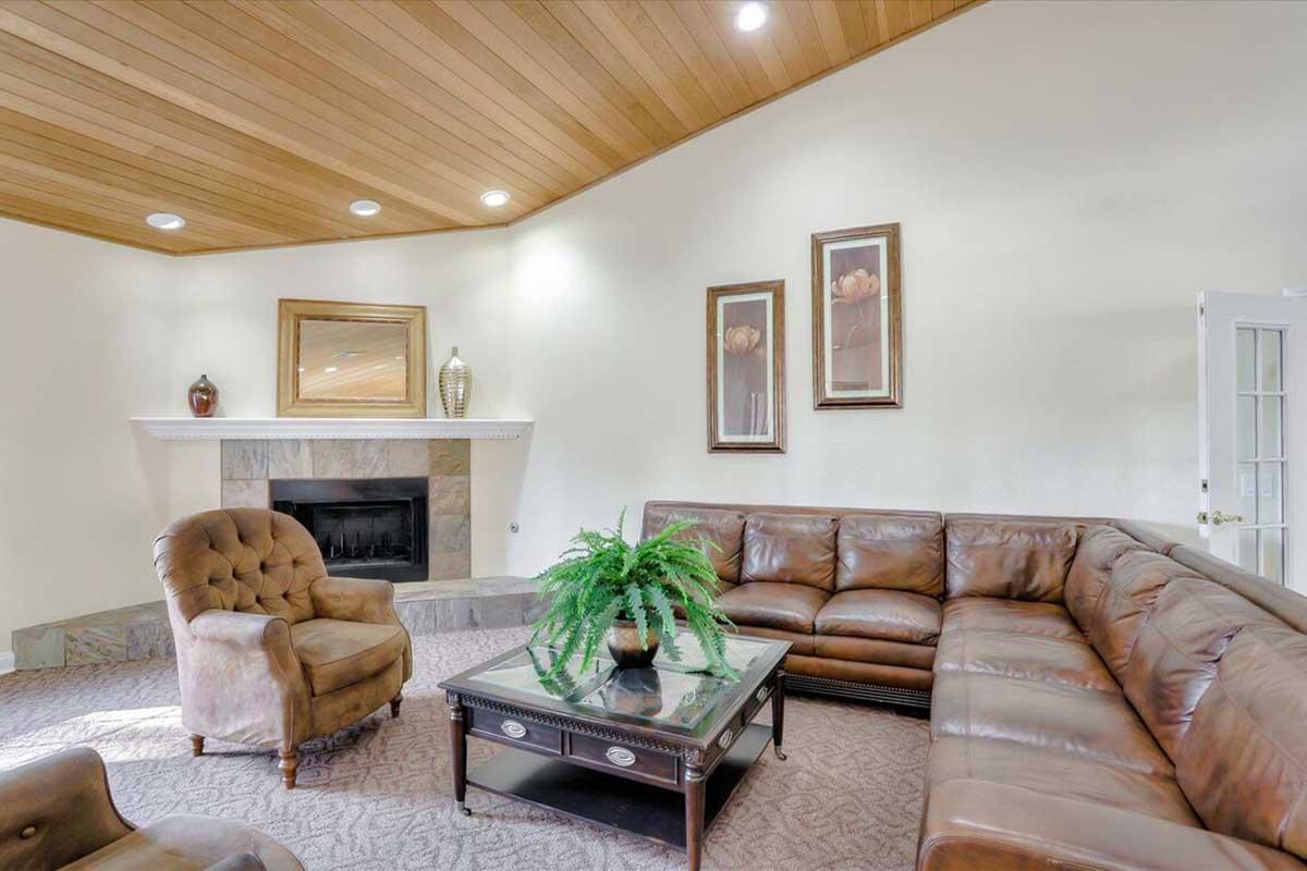 Cozy living room featuring a brown leather sectional sofa, an armchair, and a coffee table. A fireplace is framed by decorative vases and artwork on the wall. The ceiling is wooden with recessed lighting, and the floor is carpeted.
