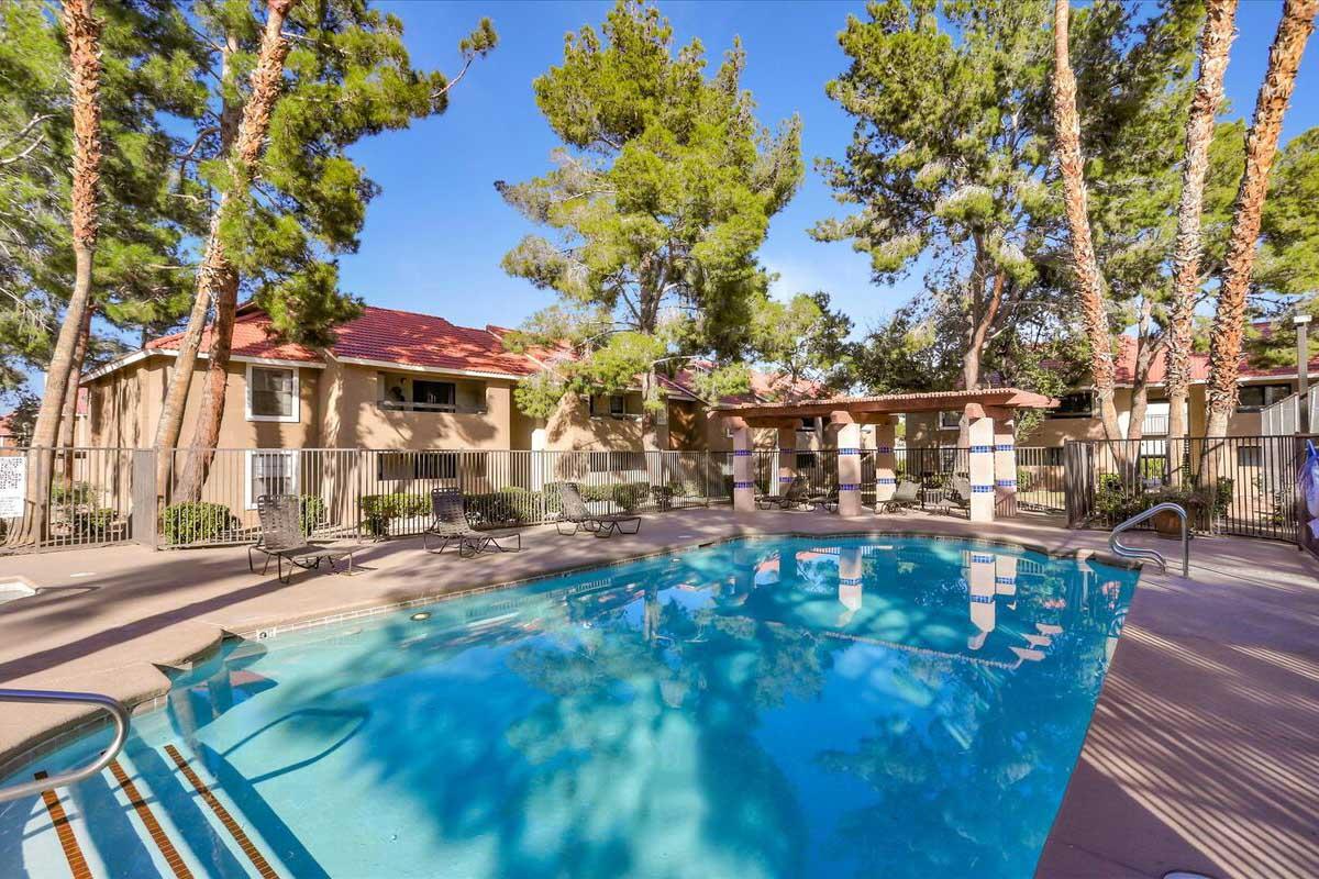 A clear swimming pool surrounded by lounge chairs and tall trees, with residential buildings in the background. The scene is well-lit and features blue skies overhead, creating a serene atmosphere for relaxation.
