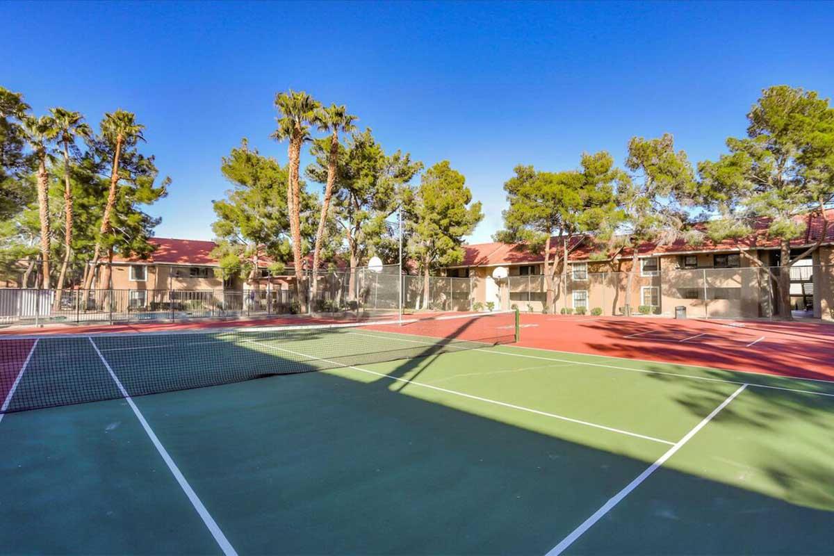 A tennis court is highlighted in the foreground, featuring a net and well-maintained surfaces. In the background, there are several tall palm trees and a residential building with red roofs, under a clear blue sky. Shadows from the trees create a pattern on the court surface.