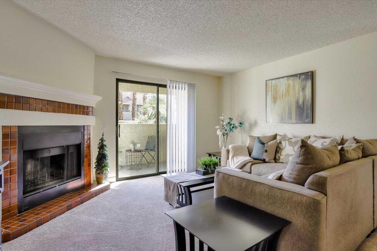 A cozy living room featuring a light-colored sofa with decorative pillows, a black coffee table, and a modern fireplace. There is a sliding glass door leading to a balcony with a small dining set. Soft lighting and a decorative plant enhance the warm and inviting atmosphere.