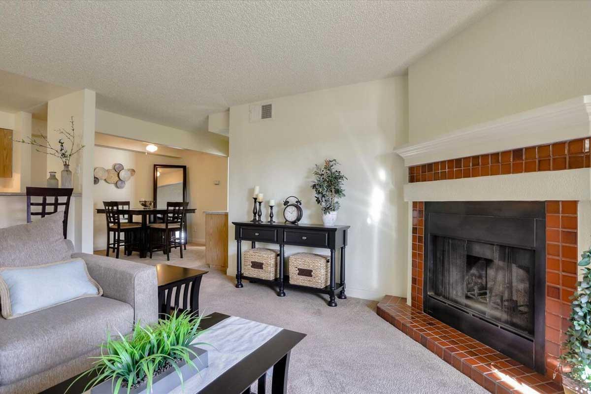 A cozy living room featuring a light grey sofa with decorative pillows, a black coffee table with a green plant, and a brick fireplace. There is a dining area in the background with a wooden table and chairs. Soft lighting and a warm color palette create an inviting atmosphere.