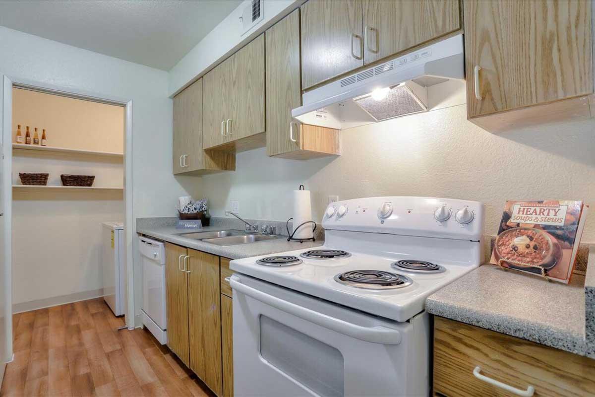A well-lit kitchen featuring wooden cabinets, a white stove with four burners, a dishwasher, and a countertop. There is a cookbook open on the counter, and a pantry door is visible in the background. The flooring is wood-like, contributing to a warm and inviting atmosphere.