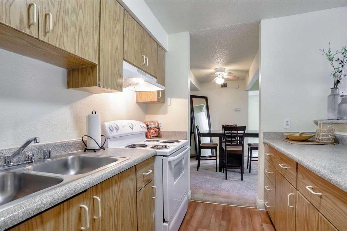 Modern kitchen with wood cabinetry and stainless steel sink. Features a white stove and hood, countertop workspace, and a dining area in the background. There’s a mirror reflecting the dining table and chairs, complemented by soft lighting and neutral-colored walls. Overall, the space is bright and inviting.