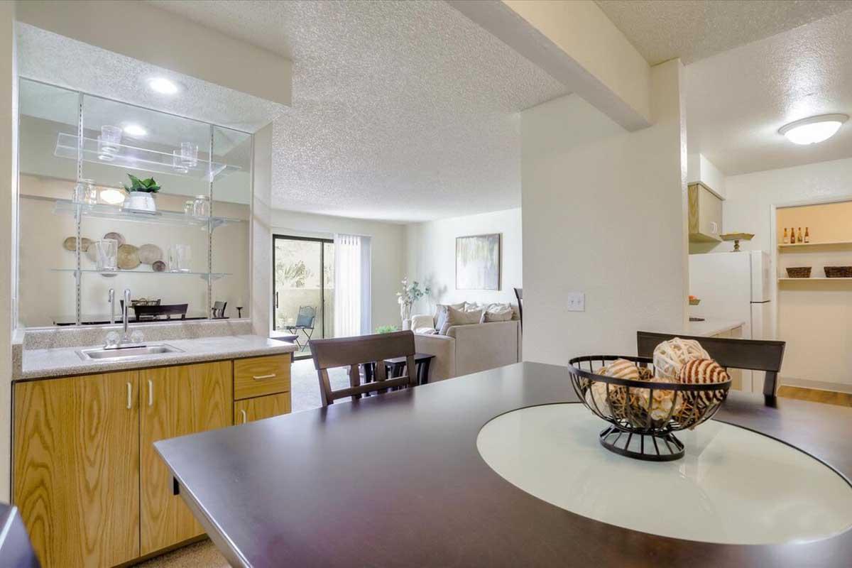 Modern kitchen and dining area featuring a wooden table with a decorative bowl, a well-organized countertop and glass shelving, leading to a living room with a couch and sliding glass doors. Bright lighting enhances the spacious and inviting atmosphere.