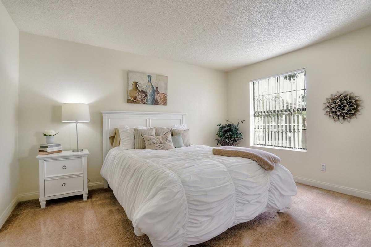 A cozy, well-lit bedroom featuring a white bed with decorative pillows, a nightstand with a lamp, and a window with blinds. The walls are painted a soft color, and there are decorative elements like artwork and a plant adding charm to the space. Soft carpeting completes the inviting atmosphere.