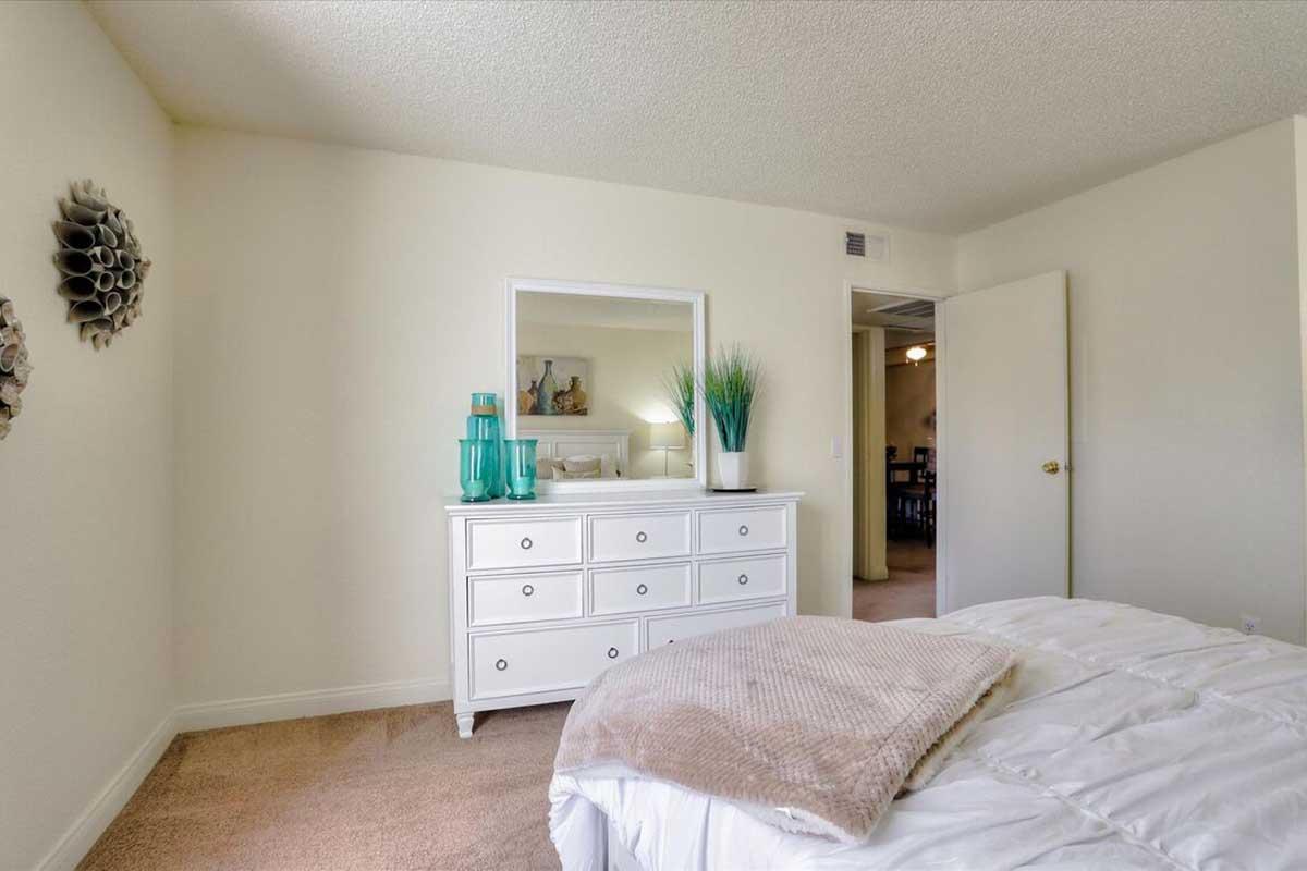 A bright and tidy bedroom featuring a white dresser with a mirror, a decorative plant in turquoise glass, and neutral-colored bedding. The walls are light-colored and there is a door leading to another room in the background. The carpet is a soft beige, enhancing the warm atmosphere of the space.