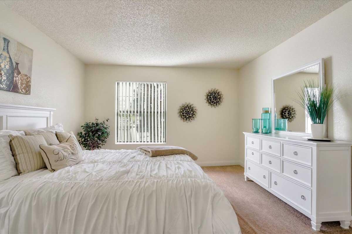 Bright and airy bedroom featuring a white bed with decorative pillows, a dresser, and a mirror. The walls are painted a soft color, and there are modern wall decorations. A window with vertical blinds allows natural light in, and a small plant adds a touch of greenery to the space.