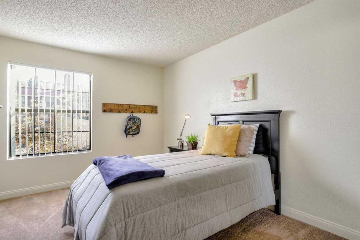 A cozy bedroom featuring a single bed with gray bedding, a small desk with a lamp and a potted plant, and a wall with a floral painting. A window with vertical blinds allows natural light to enter, and there's a wooden ruler art piece mounted on the wall. The room has neutral-colored walls and carpet.