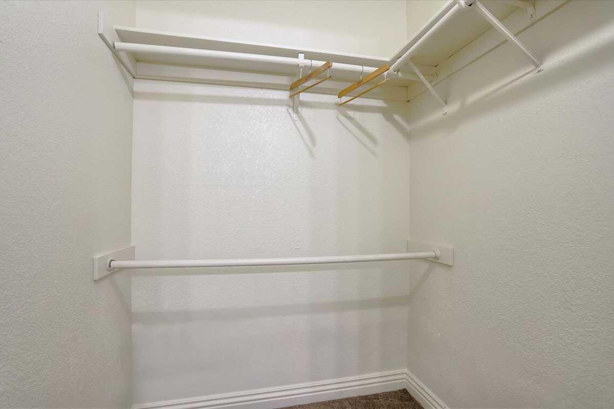 Empty closet with white walls and two horizontal clothing rods. One rod is positioned higher and is supported by two brackets, while the lower rod is longer and also mounted on brackets. The floor is covered in short brown carpet. Light is coming from the upper right, creating a bright atmosphere.