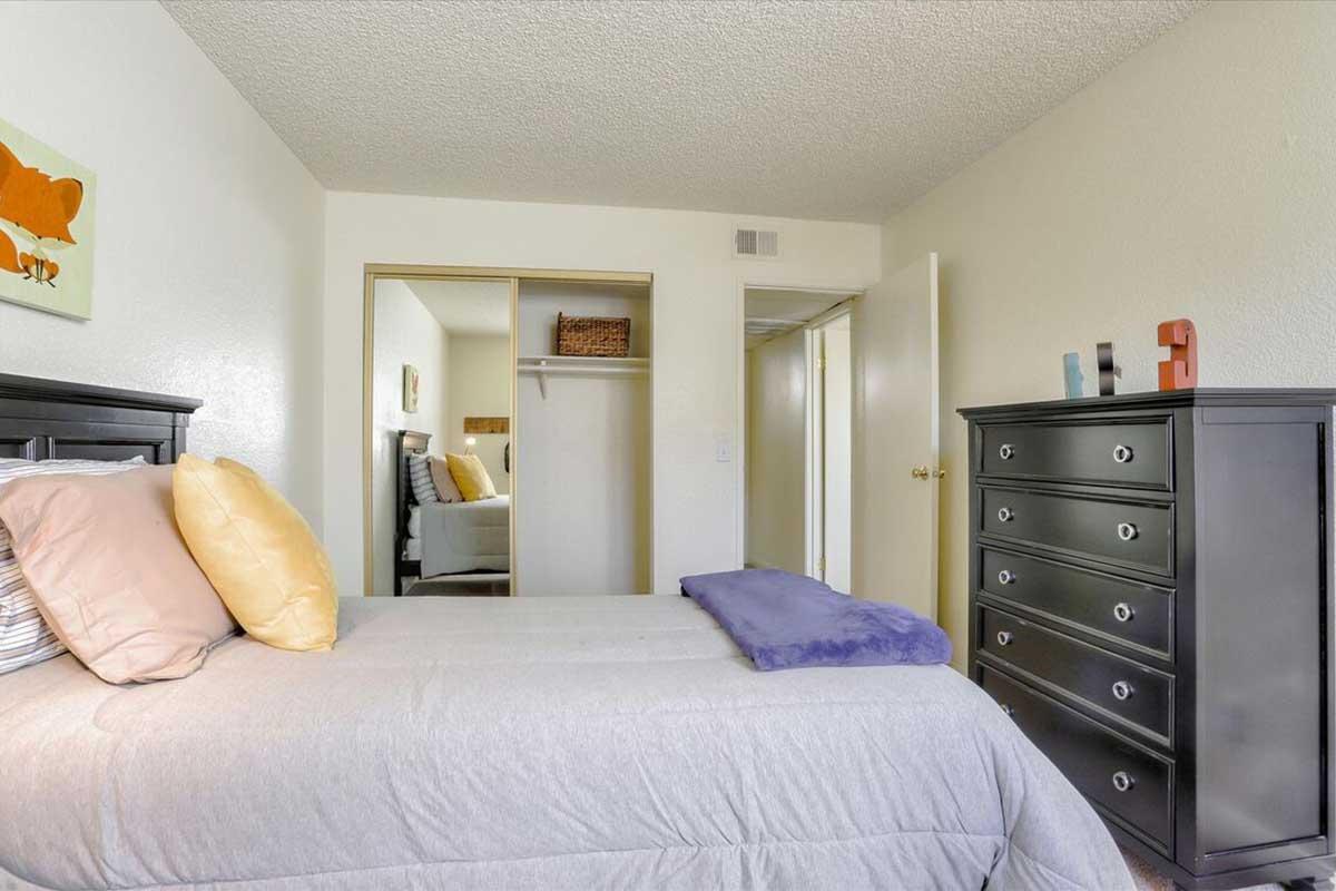 A well-lit bedroom featuring a gray bedspread, yellow and purple pillows, a dark wood dresser, and a closet with open doors. The room has light-colored walls and simple decor, creating a cozy and inviting atmosphere.