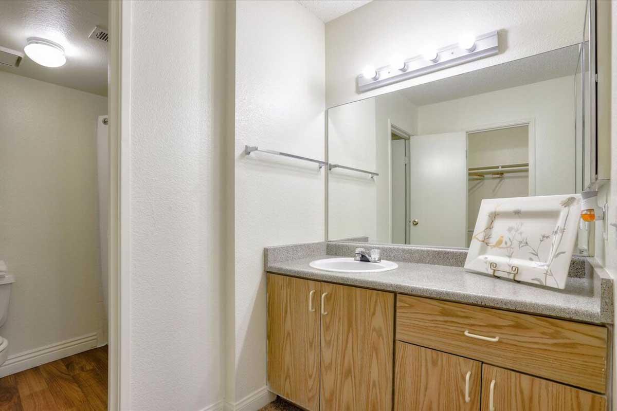 A bright bathroom featuring a small vanity with a round sink and wooden cabinets. There is a mirror above the sink, and a light fixture mounted above it. To the side, there’s a door leading to a toilet area and a small decorative framed artwork on the countertop. The floor has a wood-like finish.