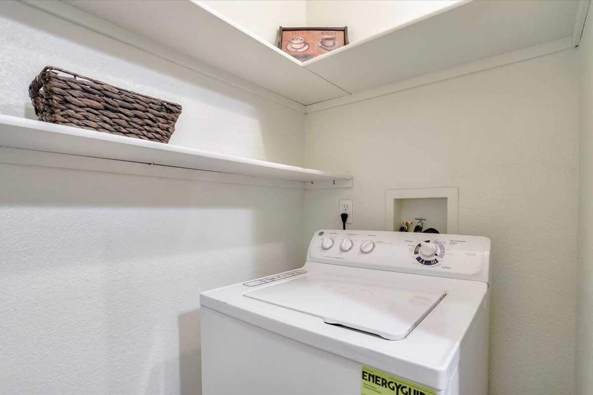 A laundry room with a white washing machine, featuring a dial and buttons on the front. Above, there are two shelves: one holds a decorative basket, and a small item is seen above the machine on the wall. The walls are painted white, and the overall space is well-lit and organized.