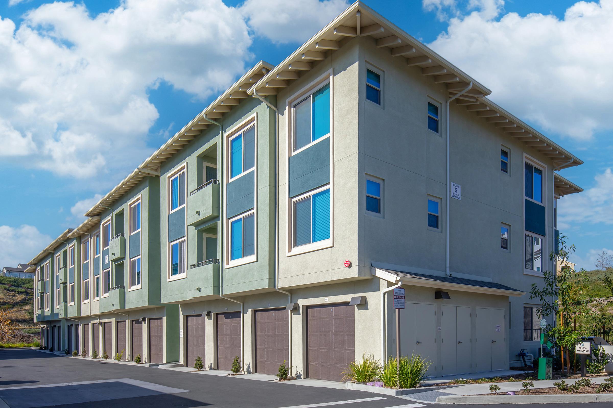 Modern multi-unit residential building with a mix of green and beige exterior. It features multiple floors with large windows and balconies. Ground-level garages are visible, and the surroundings include landscaped areas. The sky is partly cloudy, creating a bright atmosphere.