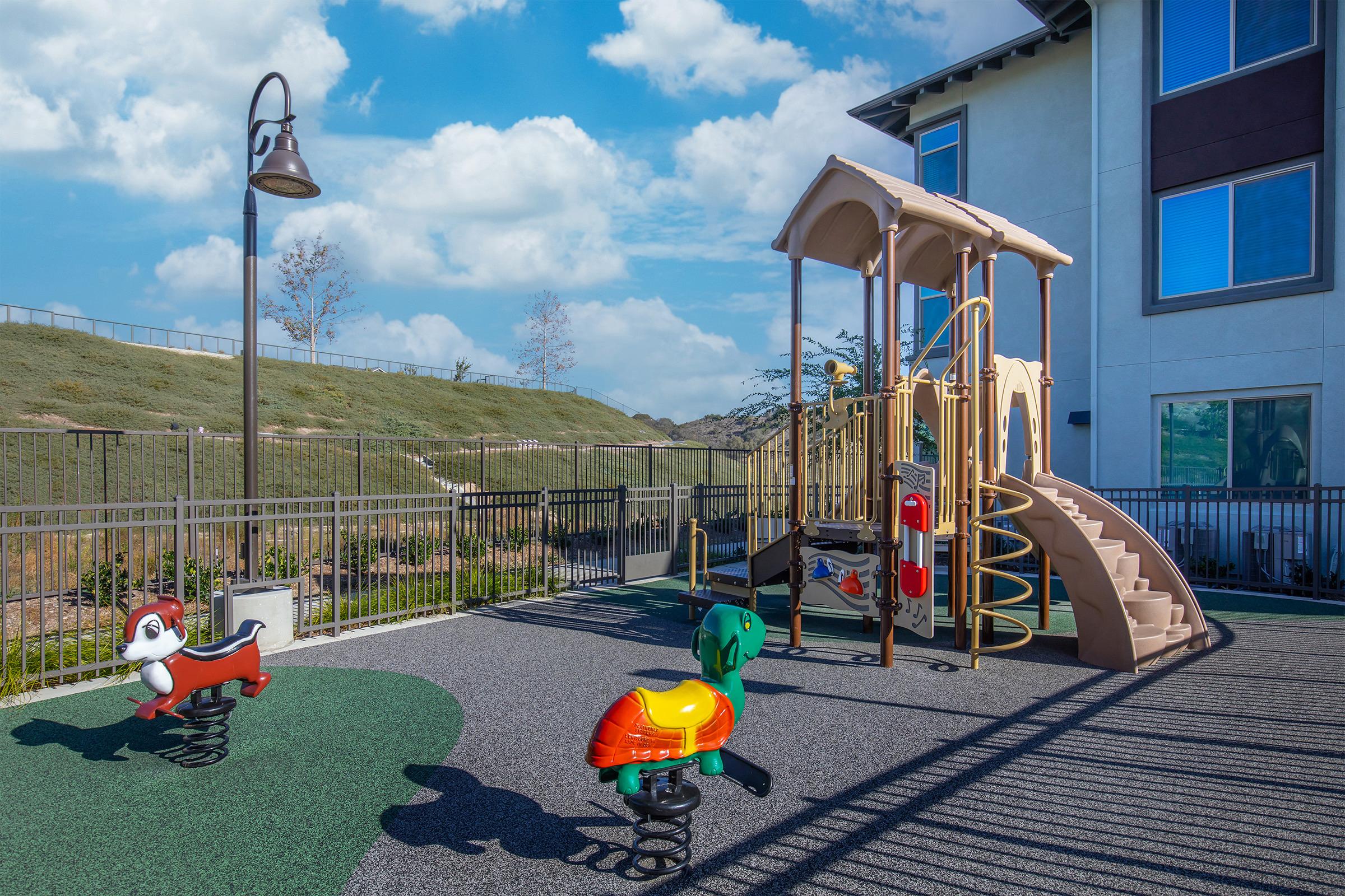 Playground area featuring a colorful slide and climbing structure, surrounded by a grassy area. Two spring riders in the foreground resemble a dog and a duck, with blue skies and fluffy clouds in the background. A fence encloses the playground, which is designed for children's recreation.