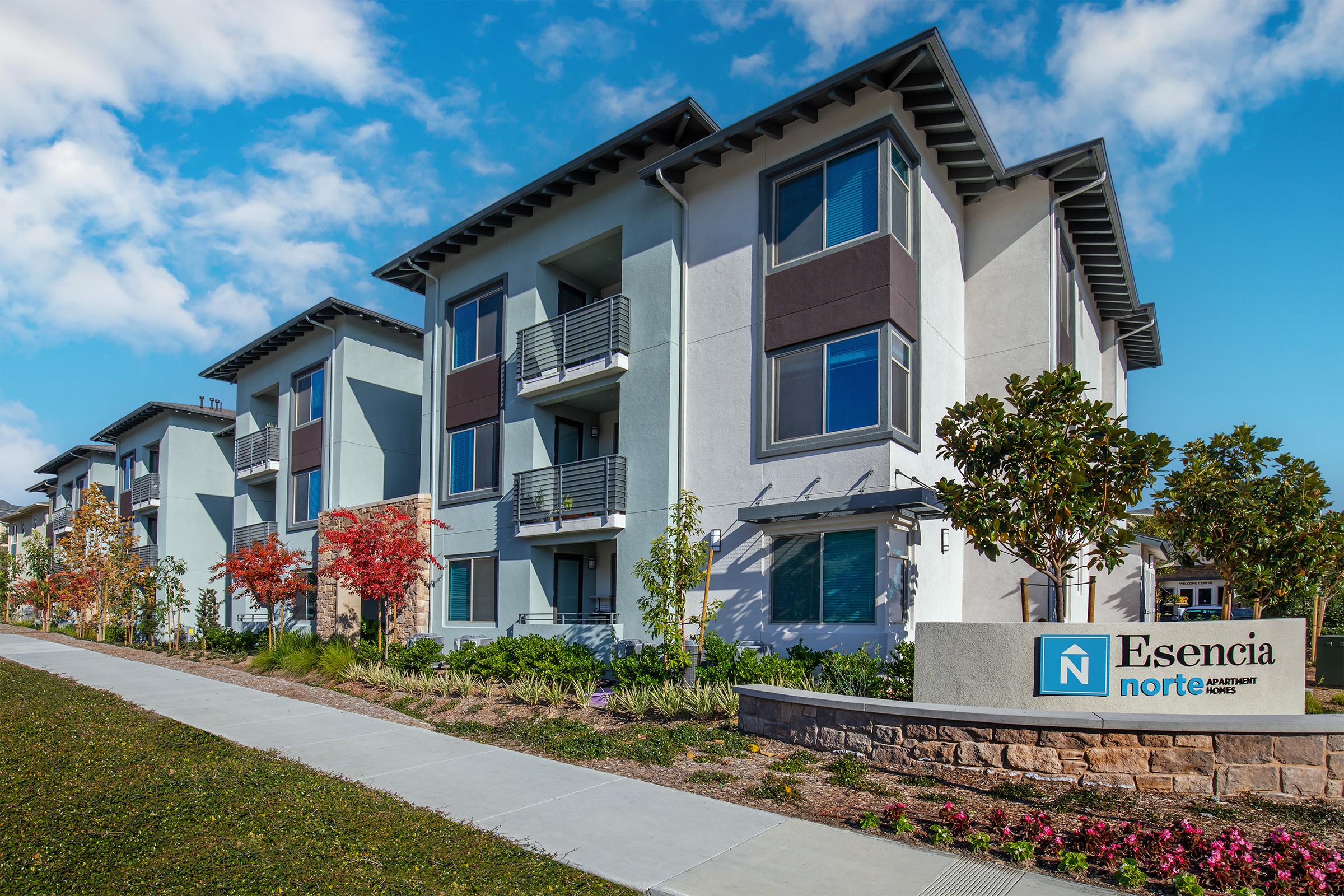 Exterior view of a modern apartment building named "Esencia Norte." The structure features a combination of light blue and gray facades, balconies, and landscaped greenery. A clear sidewalk leads to the entrance, which includes a sign displaying the apartment name. Bright skies and well-maintained surroundings enhance the inviting atmosphere.
