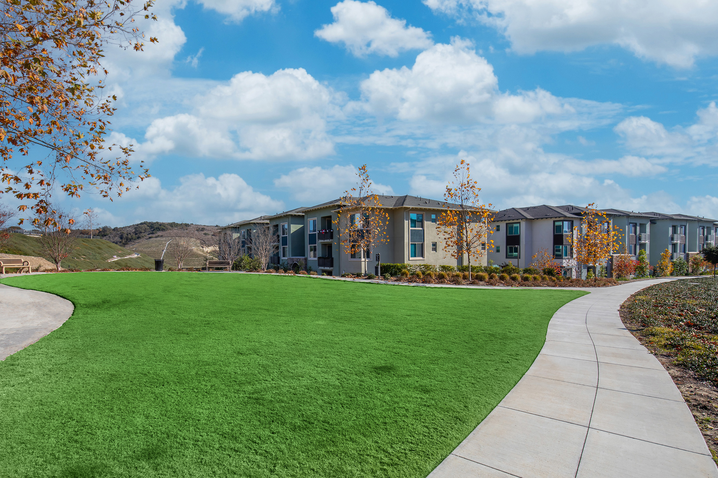 A well-maintained green lawn in the foreground, leading to a curved concrete pathway. In the background, modern apartment buildings with balconies are visible, set against a backdrop of rolling hills and a bright blue sky with fluffy clouds. Autumn leaves can be seen on trees lining the path.