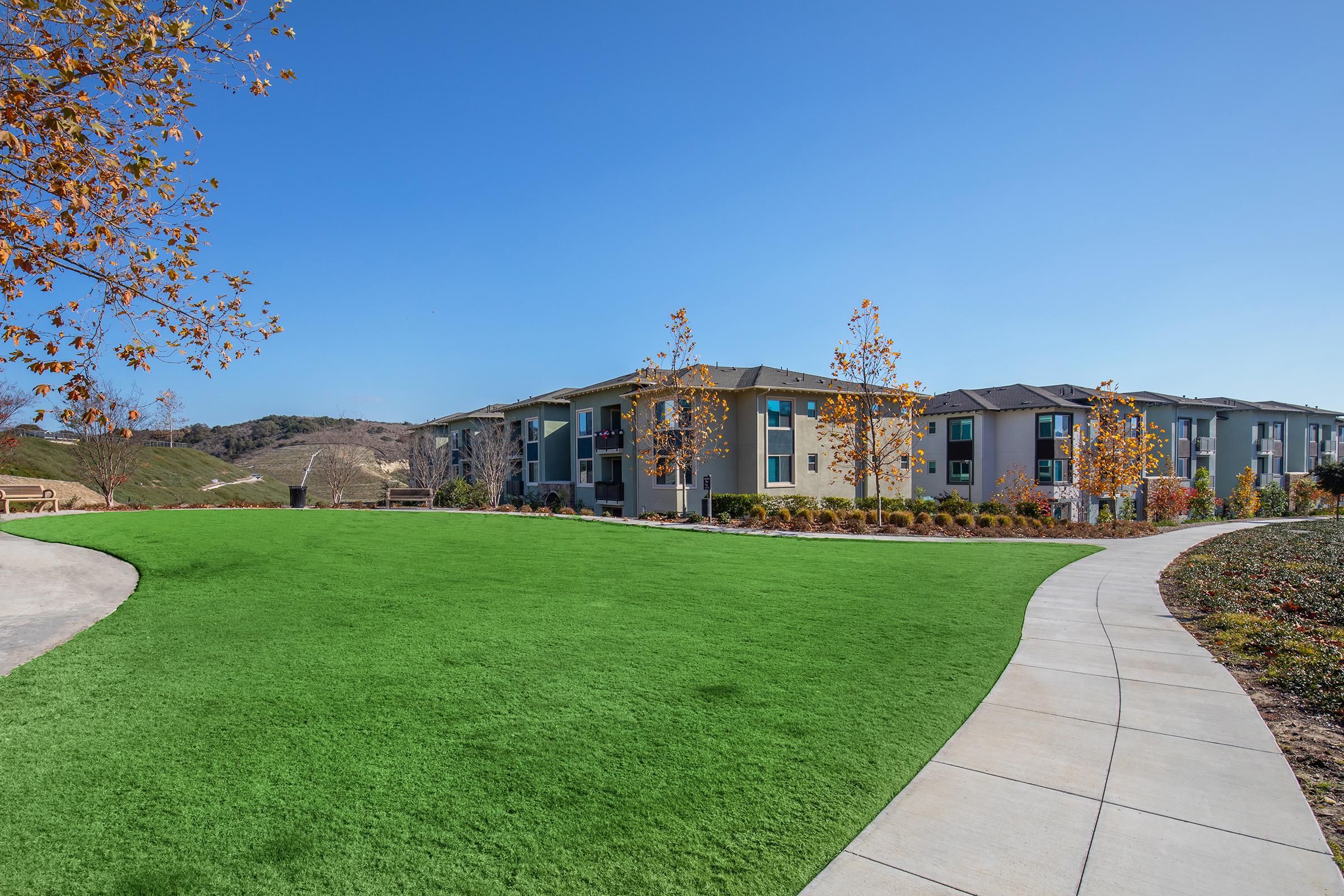 A vibrant green lawn surrounds modern apartment buildings, set against a clear blue sky. Trees with autumn leaves add a pop of color, and a paved pathway curves through the landscape, leading to the entrance of the residences. The hillside in the background suggests a peaceful, suburban setting.