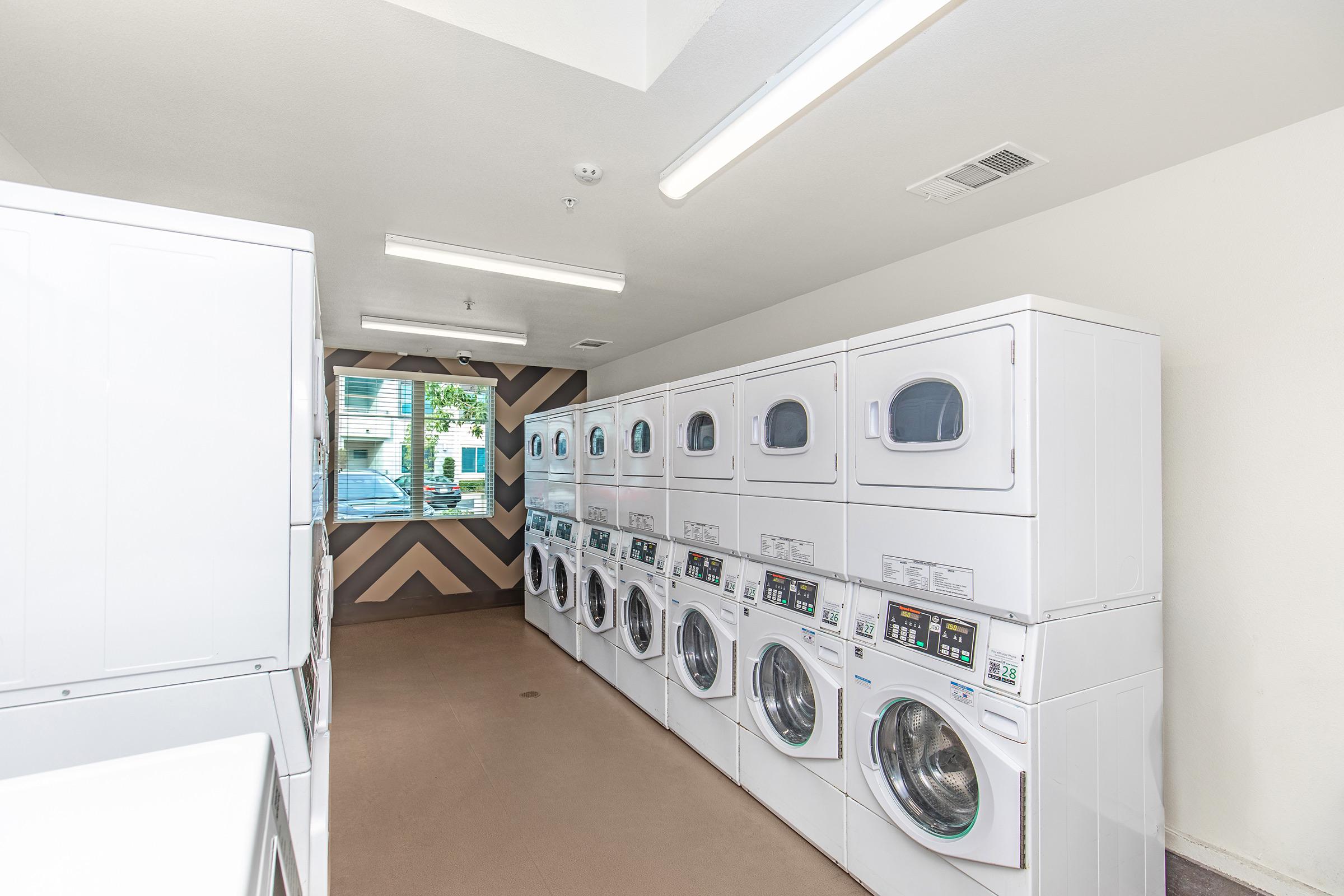A bright and clean laundry room featuring stacked washing machines and dryers along one wall. The machines have clear windows, and there are utility hooks on the ceiling. This room has a modern design, with light walls and a geometric pattern on the opposite wall, creating an inviting atmosphere for laundry tasks.