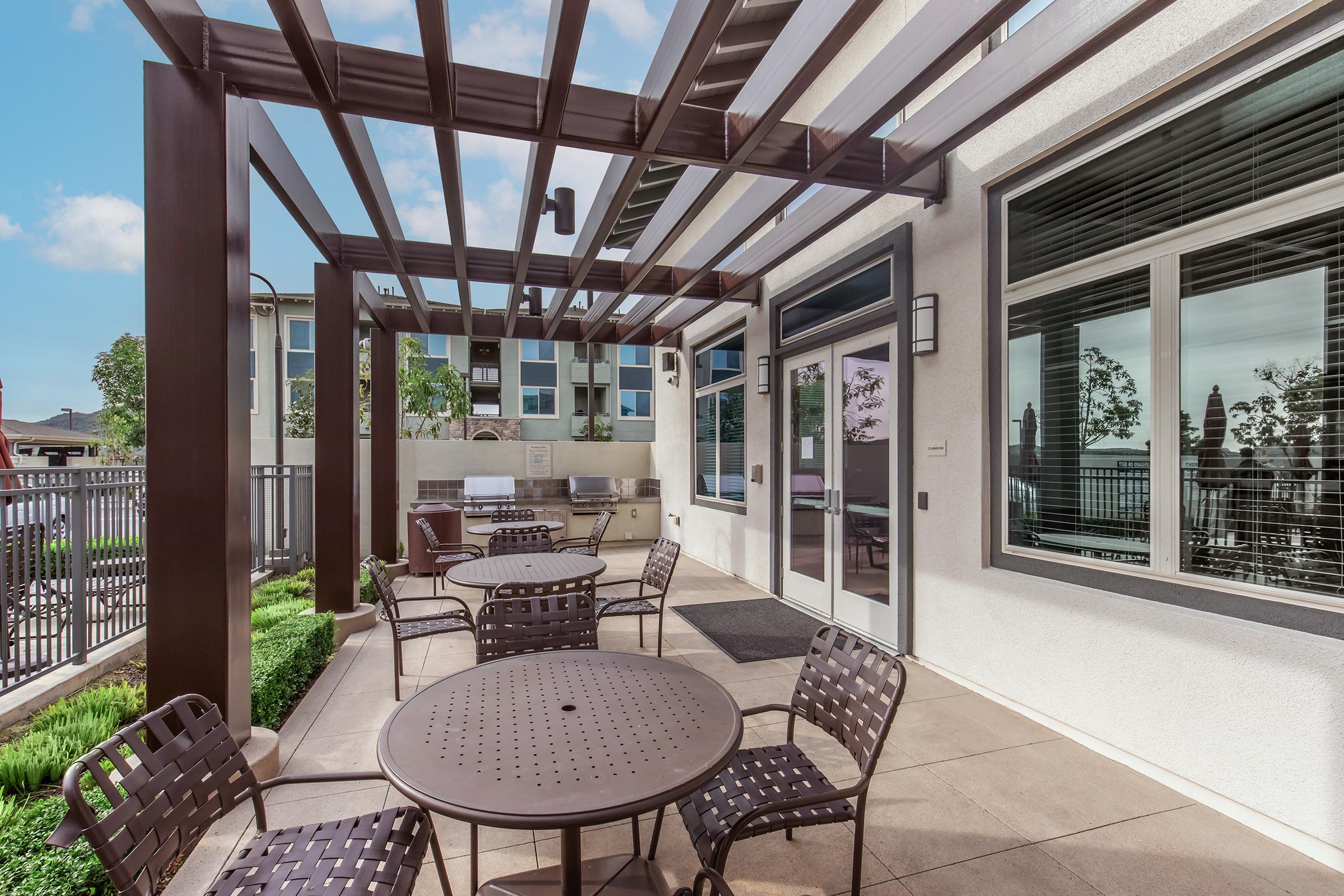 A modern outdoor patio area featuring several circular tables with chairs, shaded by a wooden pergola. There are barbecuing facilities in the background and large windows offering views of surrounding greenery. The space is well-lit and inviting for gatherings or relaxation.